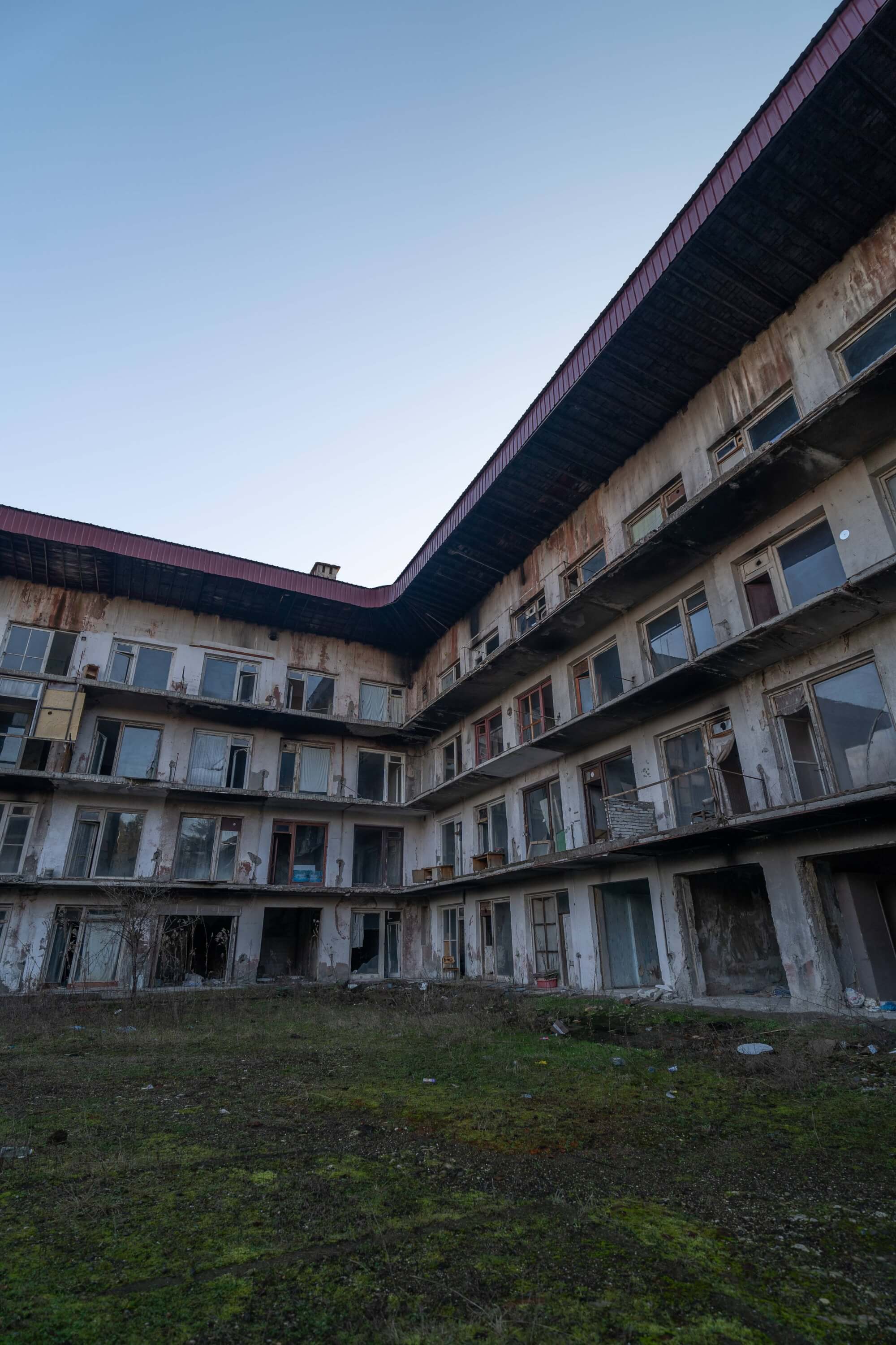 A view of a decrepit multi-story building with broken windows surrounding a grassy courtyard, under a clear sky.