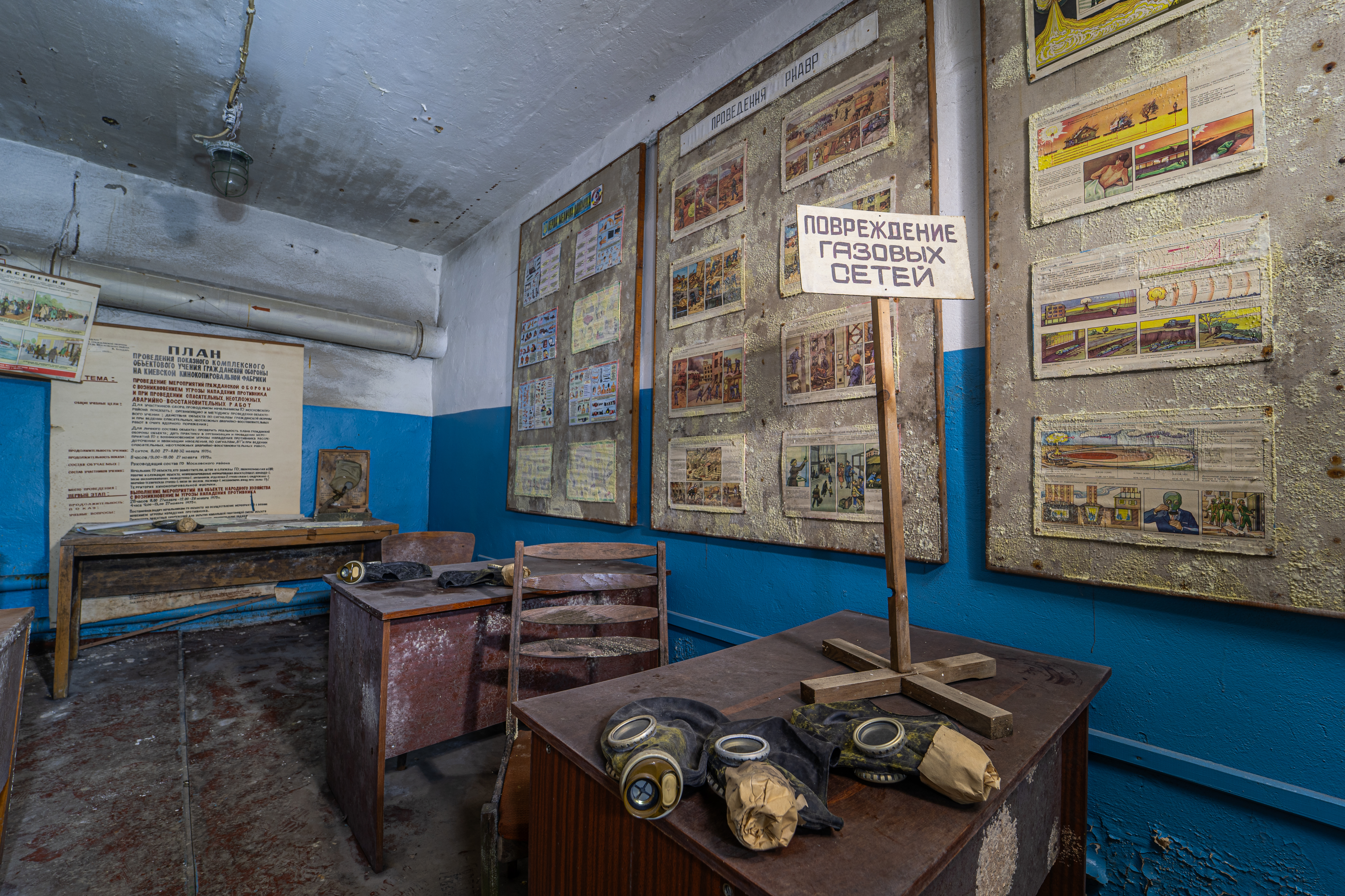 An abandoned emergency preparedness training room with wooden desks and gas masks. The walls are adorned with faded safety posters, and a sign about gas network damage is displayed prominently.