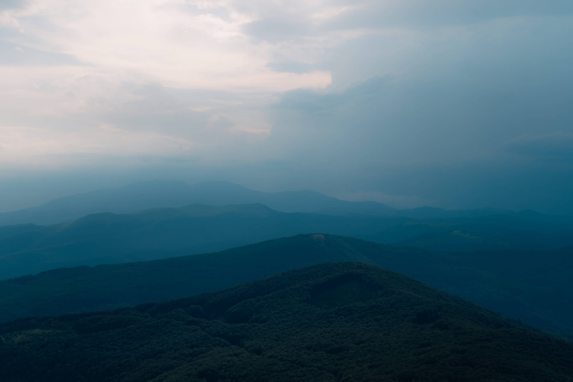 A panoramic view of misty mountains under a cloudy sky, featuring layered hills and dense greenery fading into the background.