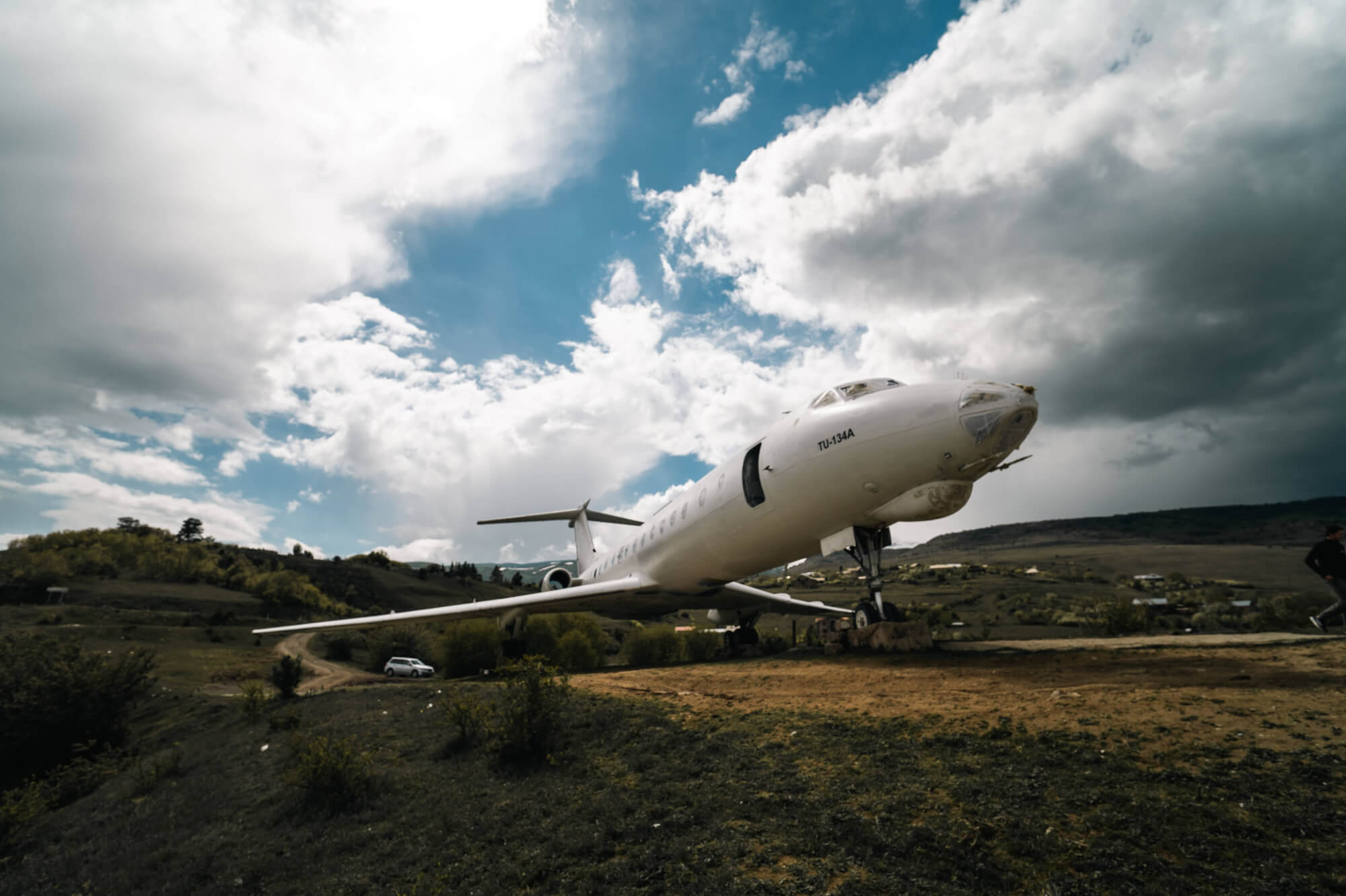 A white airplane on a hillside with a cloudy sky in the background. There is a green landscape around it, with a car and a person visible in the distance.
