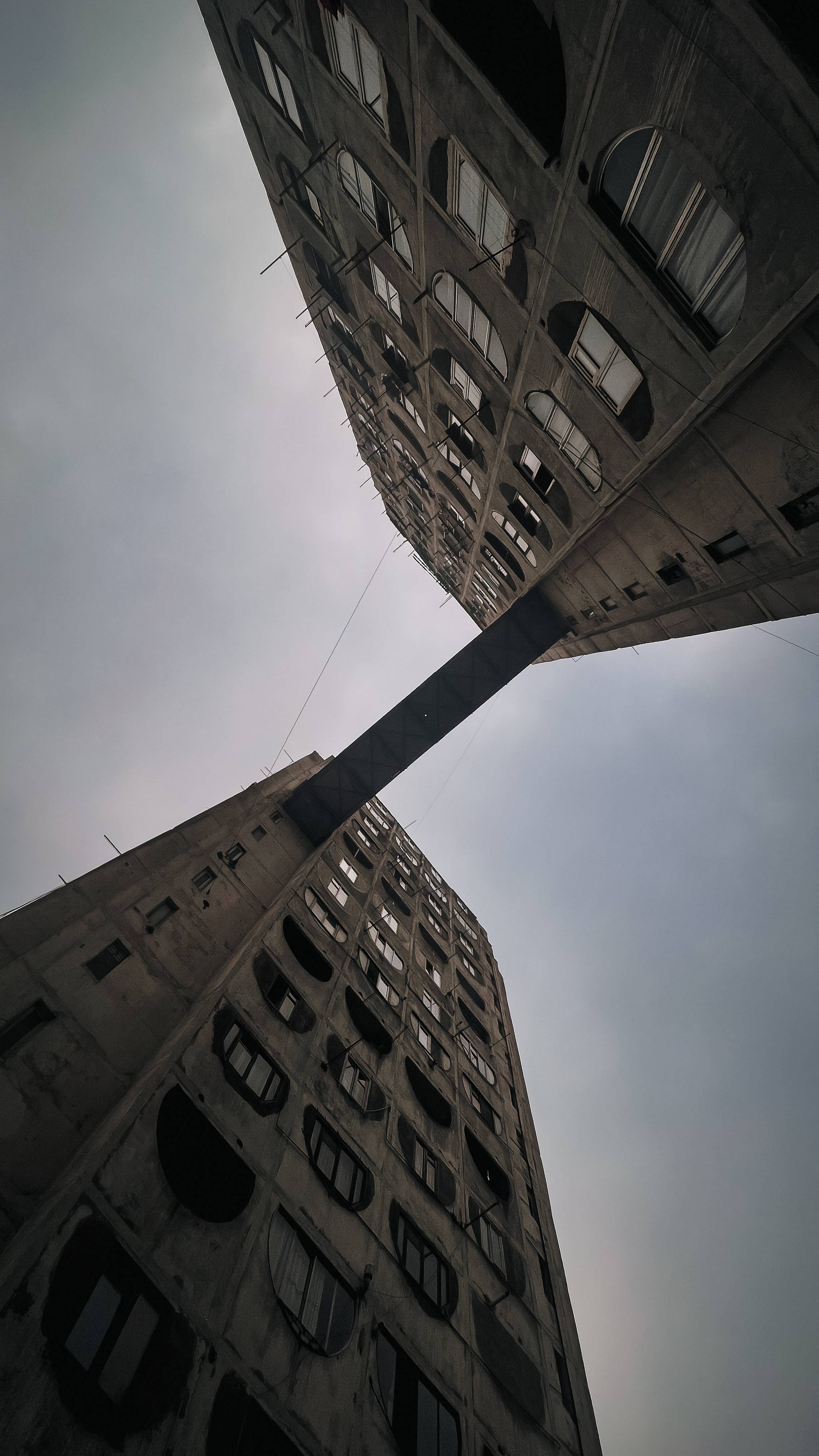 A low-angle view of two tall, decaying concrete buildings with large circular windows, converging at the top with a metal beam bridge connecting them across a cloudy sky.