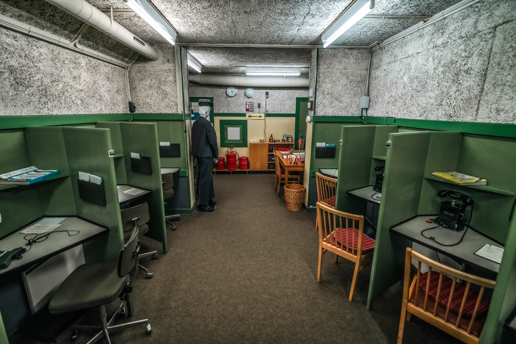 A view of an empty office space with green cubicles, a man in a suit standing against the wall, old-fashioned telephones on desks, and a wooden table with office supplies.