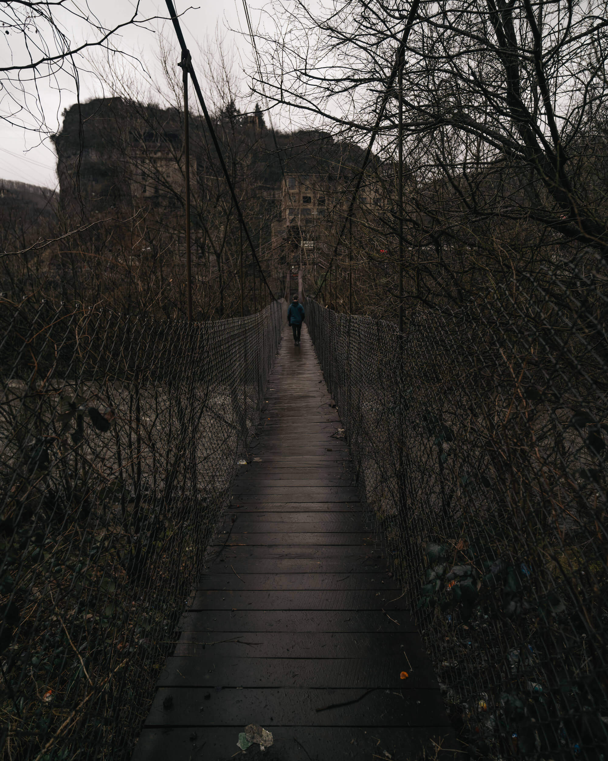 A person in a blue jacket walking on a wooden bridge surrounded by bare trees and an abandoned building in the background under a cloudy sky.