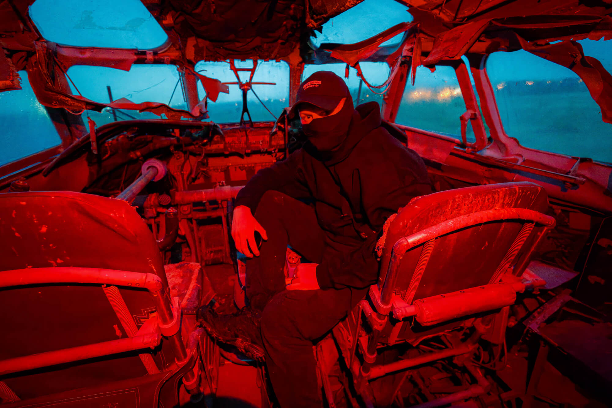 An abandoned aircraft cockpit with a person in dark clothing and a mask sitting inside, surrounded by worn pilot seats and control equipment, bathed in red and blue light.