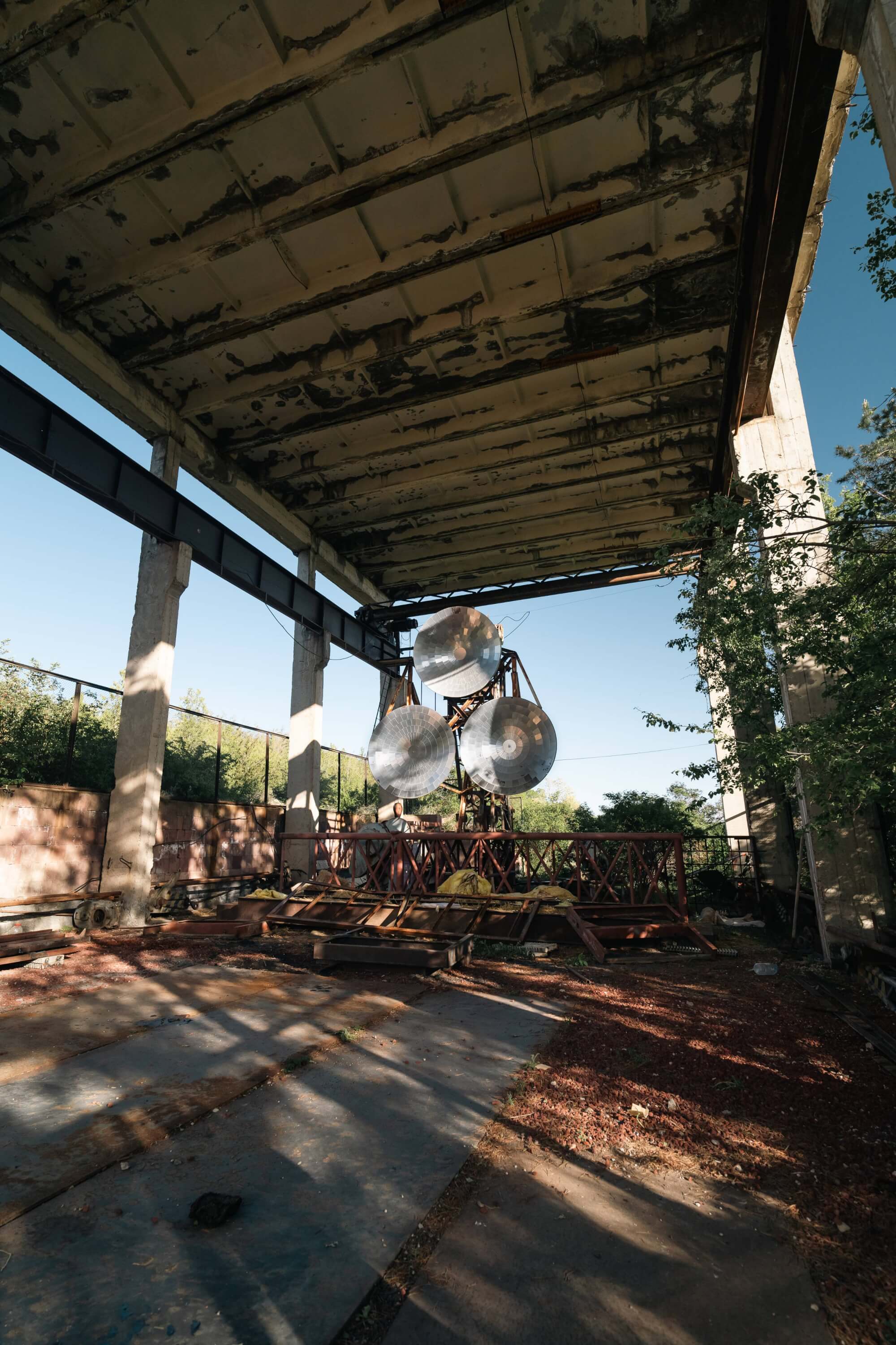 An abandoned industrial interior with a rusted ceiling, large metallic discs hanging from a structure, and greenery around the edges.