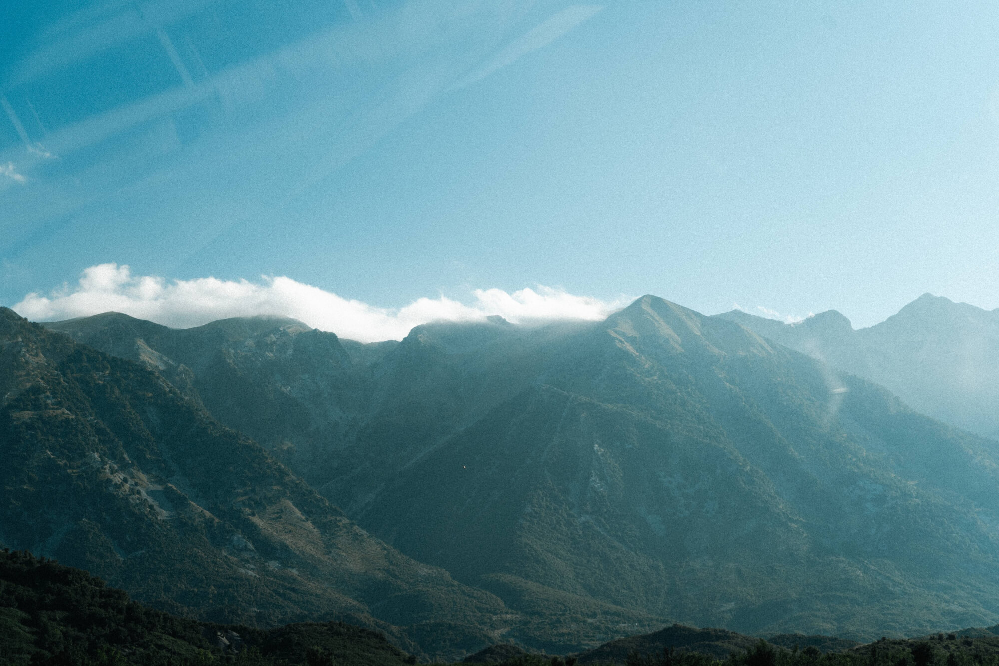 A panoramic view of mountains with lush greenery in the foreground and rocky peaks in the background, under a clear sky with scattered clouds.