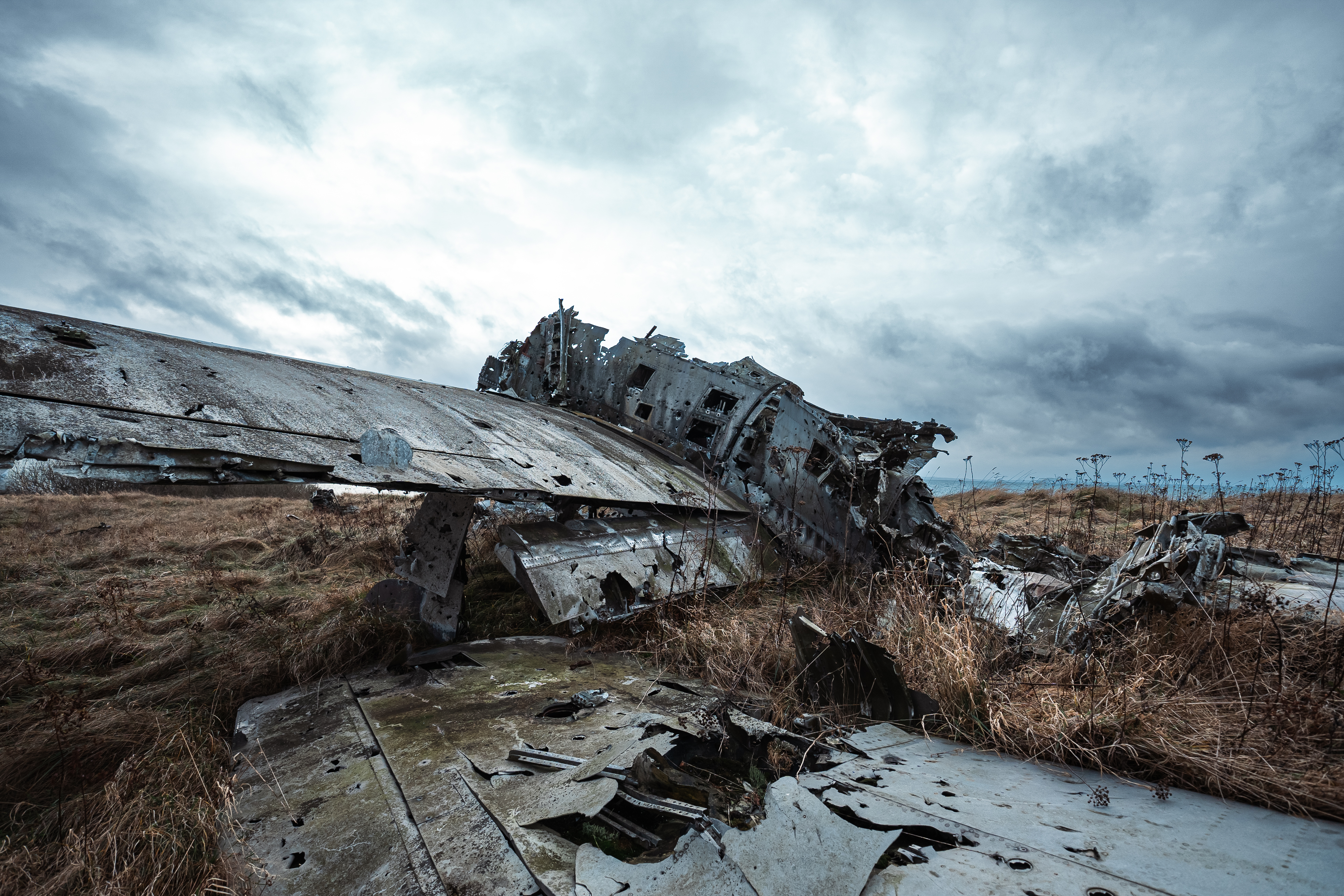 A wrecked airplane fuselage lies on its side in an open field, surrounded by dry grass and sparse vegetation. Dark clouds fill the sky above, creating a gloomy atmosphere.