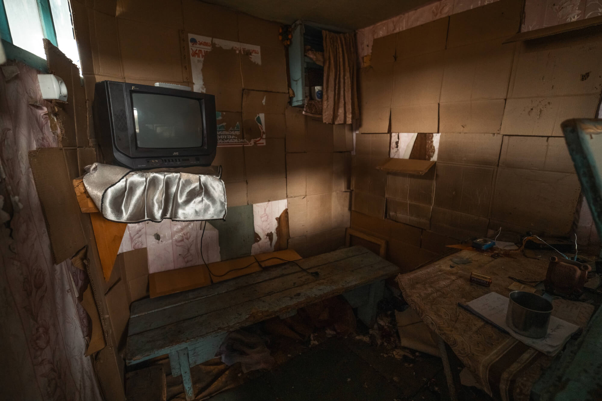 A small, cluttered room with cardboard walls, a vintage television on a wooden shelf, a table with various objects including a cup and papers, and a dimly lit atmosphere.