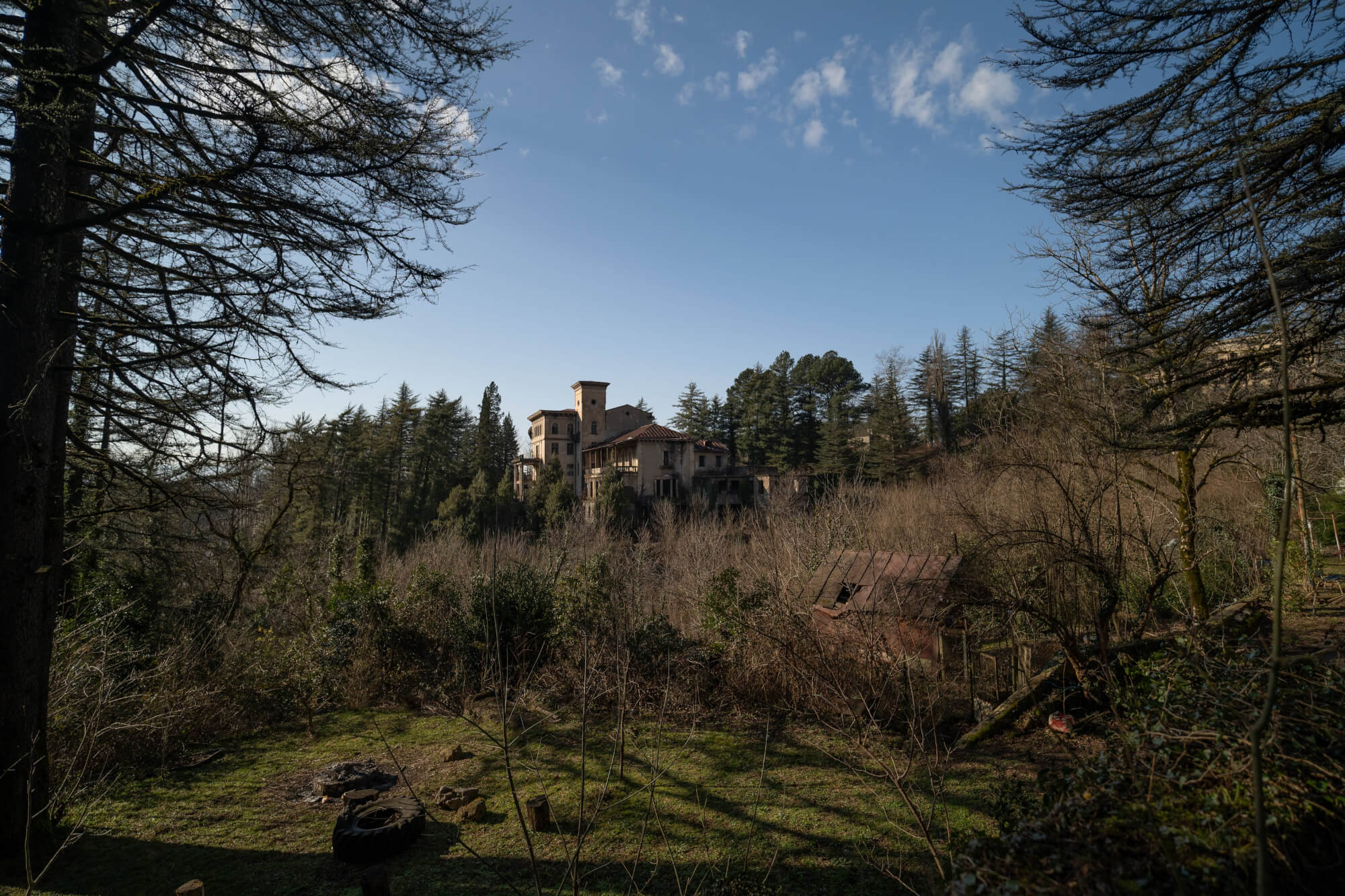 A partially abandoned castle surrounded by dense trees, with an old shed and firepit in a grassy clearing in the foreground.