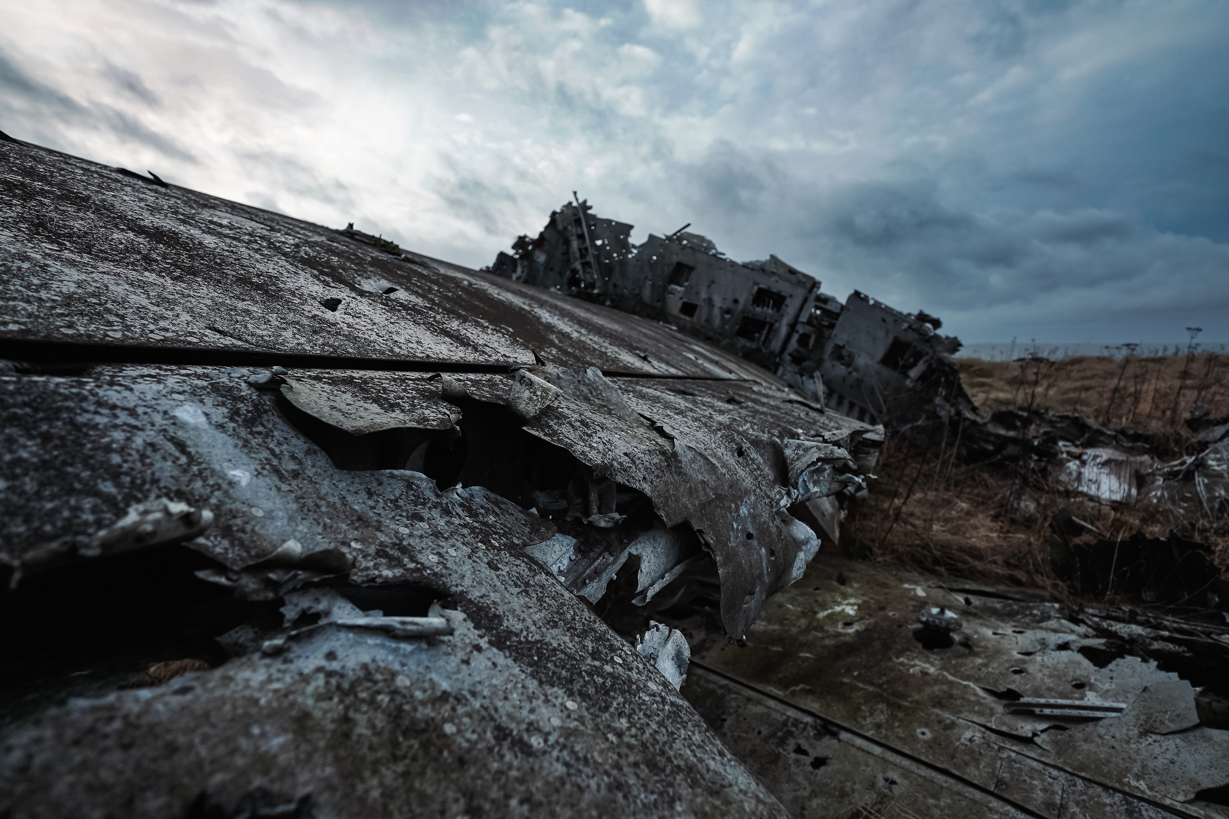 Close-up view of a decaying metallic surface with rust and peeling paint, with parts of an abandoned aircraft visible in the background under a cloudy sky.