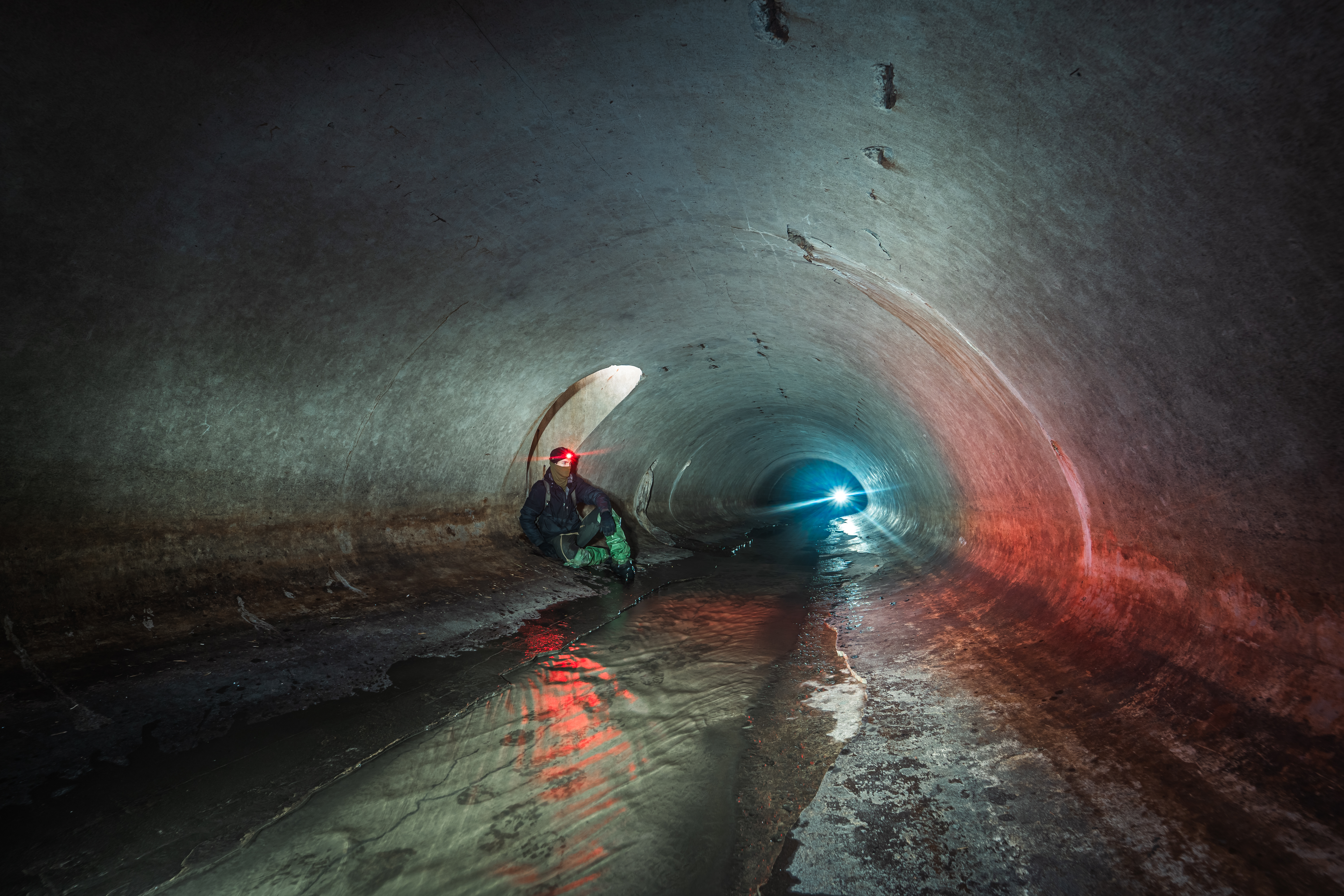 A person sitting in a dark underground tunnel, illuminated by colorful lights, with water reflecting on the ground.