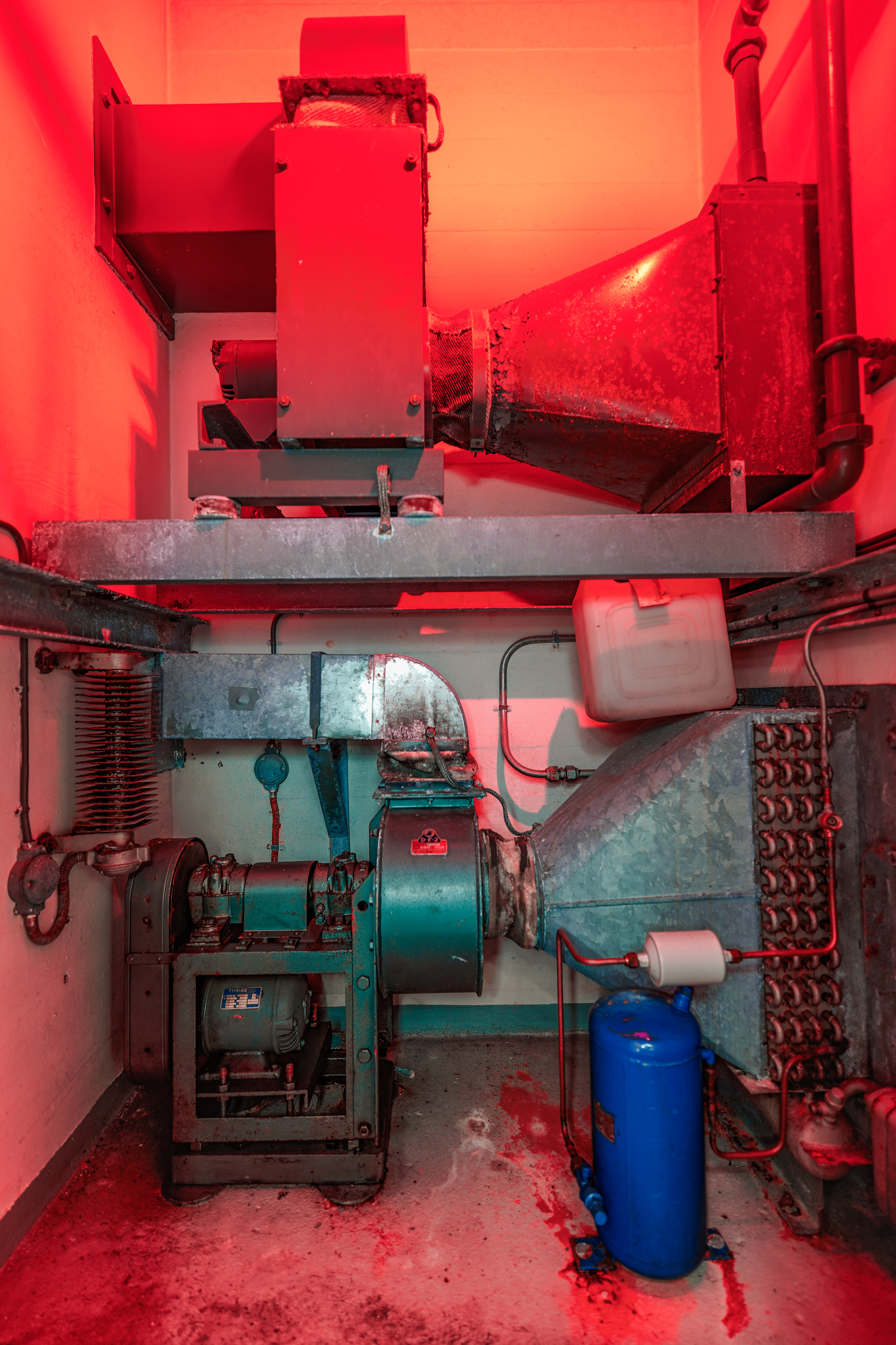 An industrial machine setup featuring a large fan, pipes, and a blue tank, illuminated by red lighting creating a dramatic atmosphere.