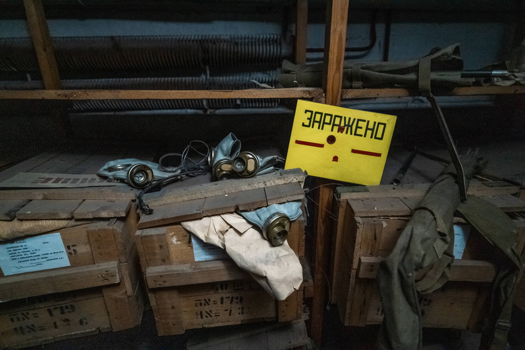 A dimly lit room displaying wooden crates with some partially open, a yellow warning sign saying 'ЗАРАЖЕНО', and gas masks scattered on top.
