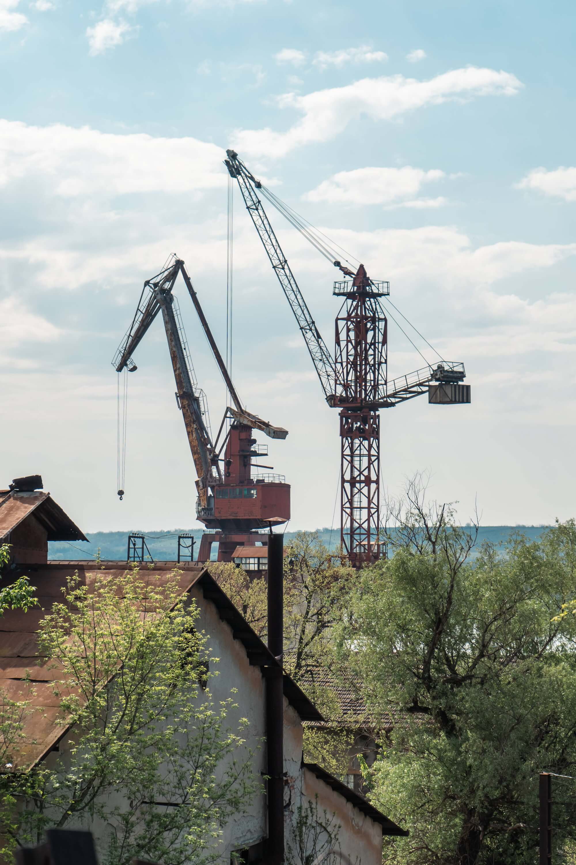 An industrial landscape featuring two large cranes against a cloudy sky, surrounded by old rooftops and greenery.