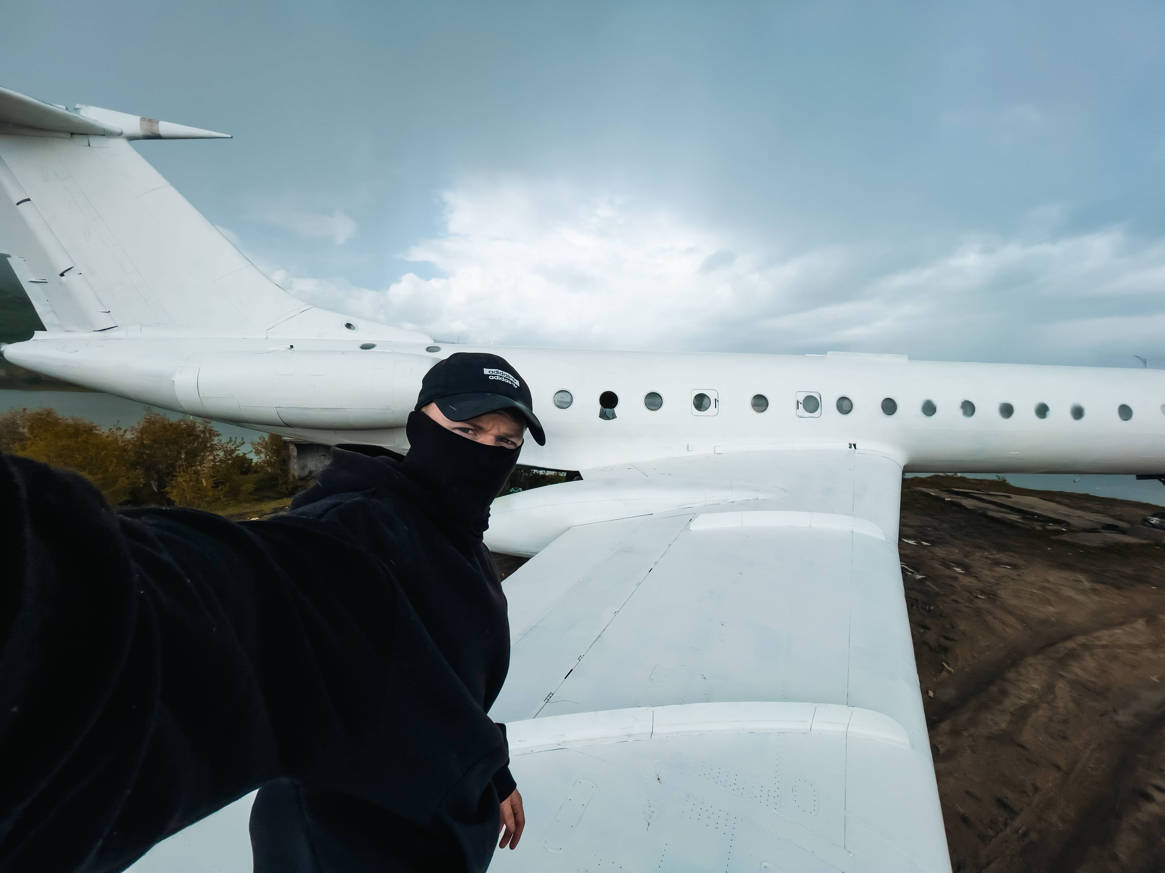 A person wearing a mask and cap stands on the wing of a white airplane against a cloudy sky, with a landscape featuring trees in the background.