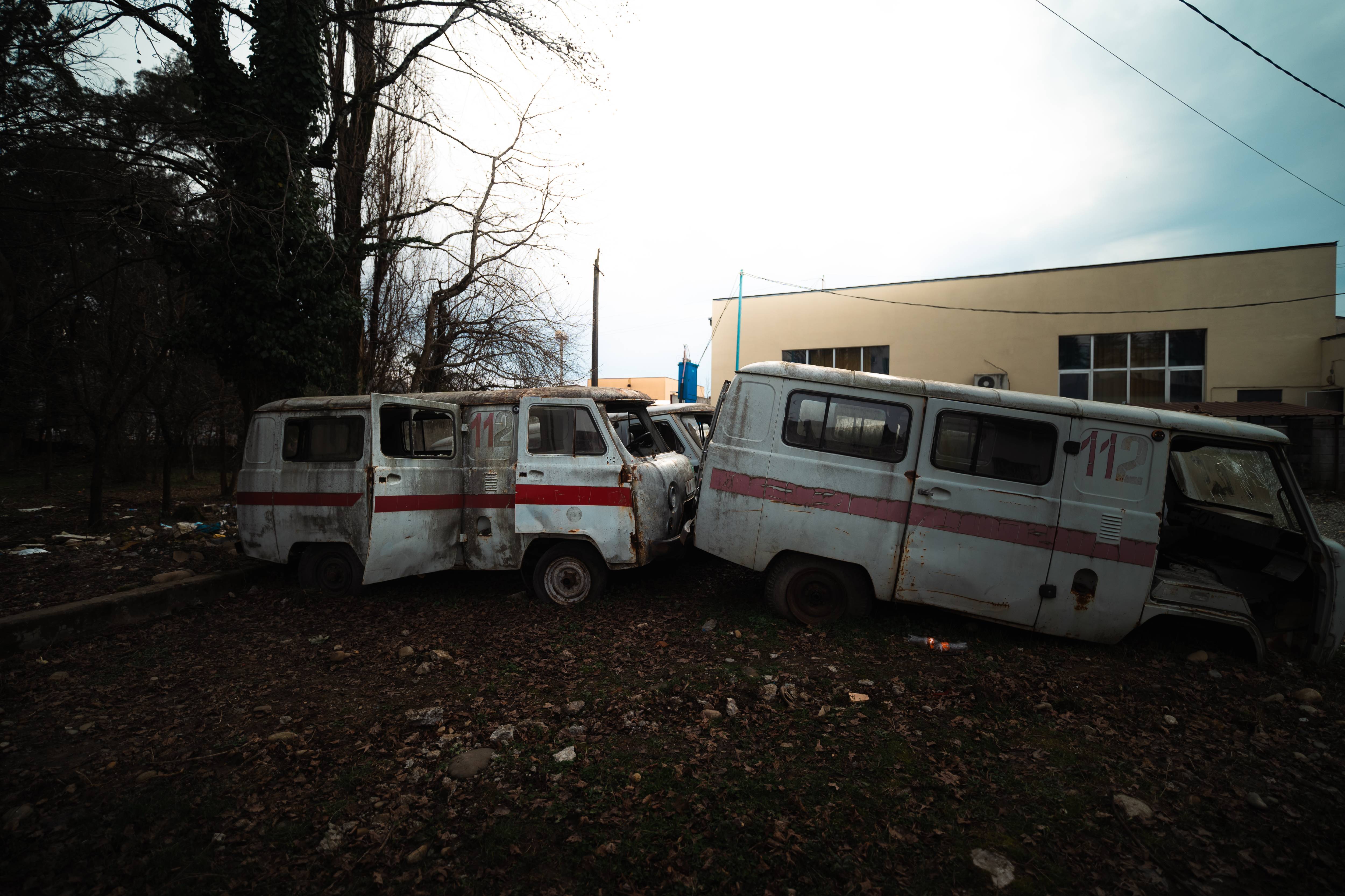 A cluster of abandoned, rusty ambulances with faded white and red colors, partially obscured by surrounding trees and scattered leaves, set against an overcast sky and a backdrop of an industrial building.