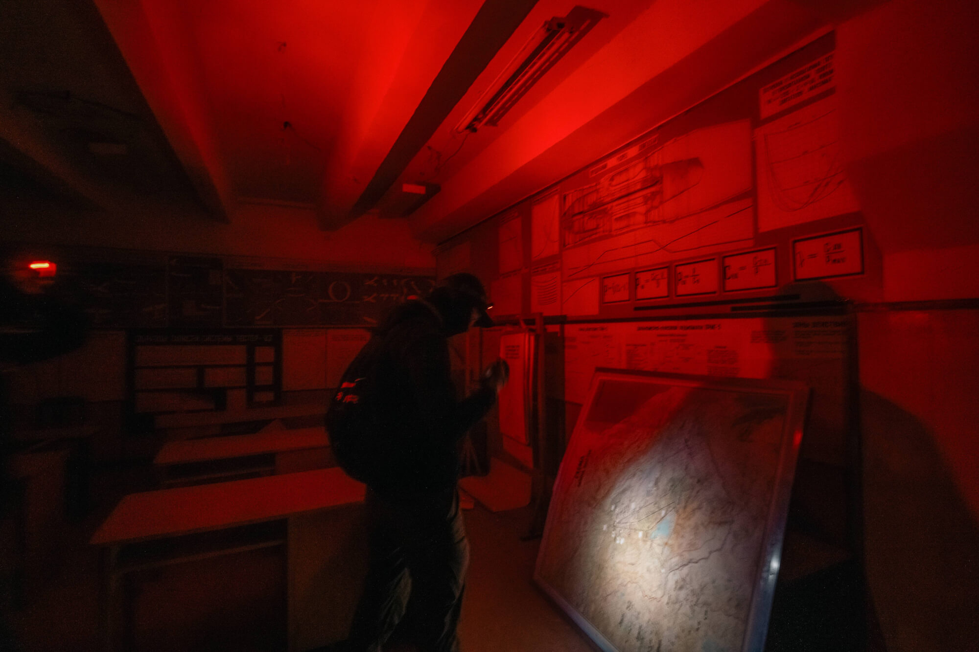 A dark room illuminated with red light, featuring a person inspecting a map on a table, with shadowy furniture around.