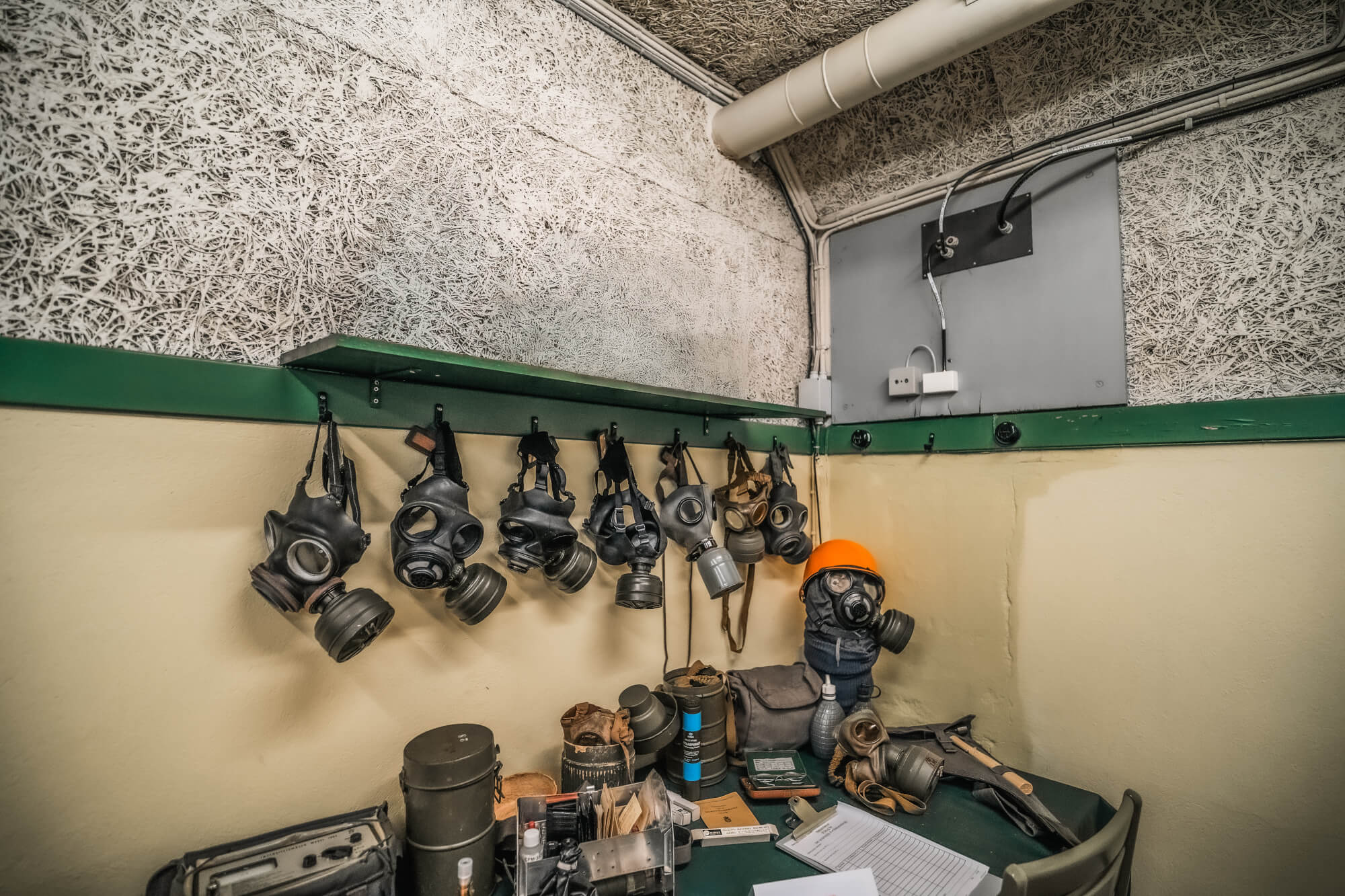 A corner of a room displaying eight gas masks hanging on a wall, an orange helmet on a mannequin, and a table cluttered with tools and supplies, set against textured walls.