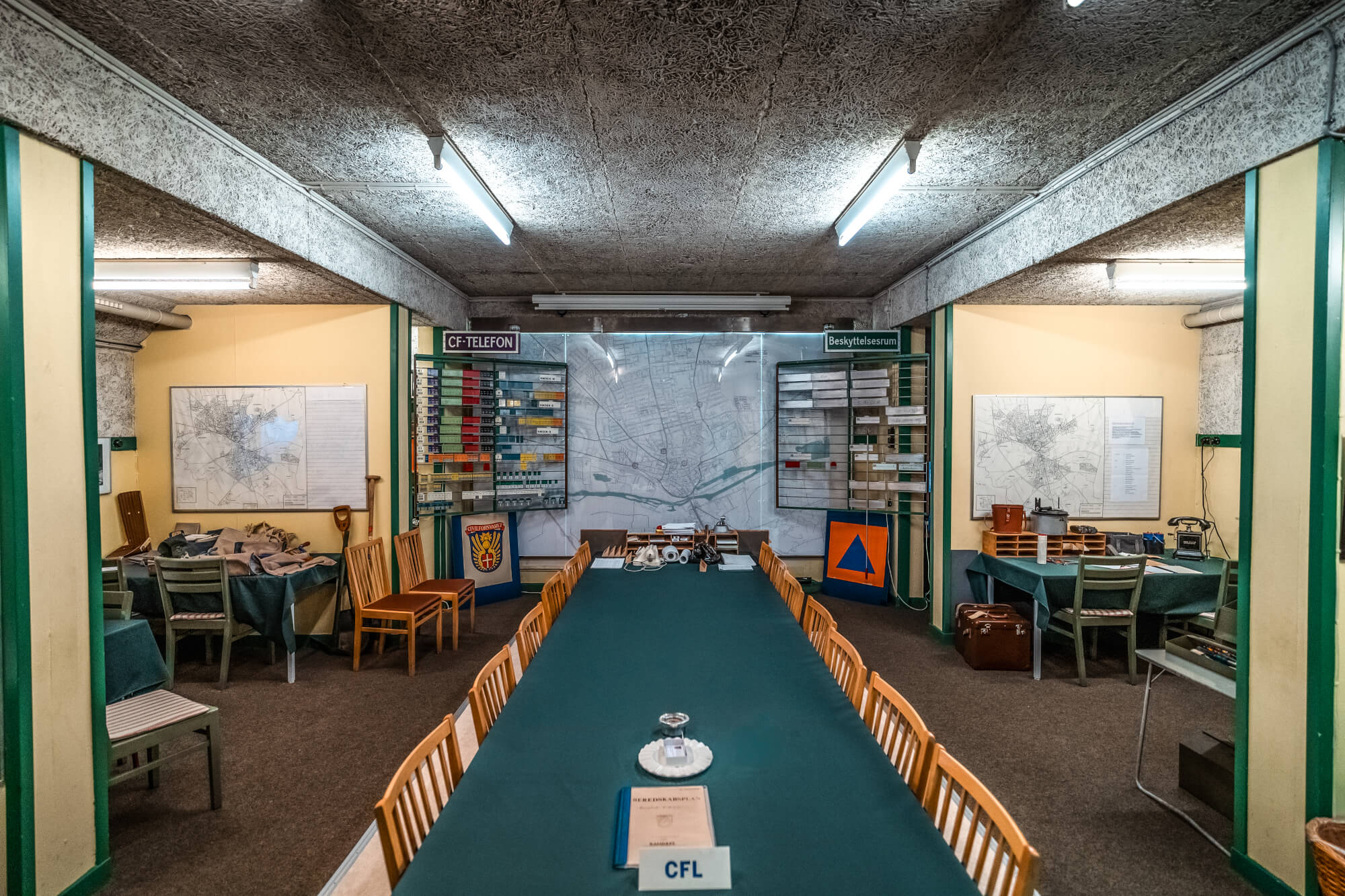 A historical command center with a long green table, wooden chairs, and walls adorned with maps and communication equipment, illuminated by fluorescent lights.