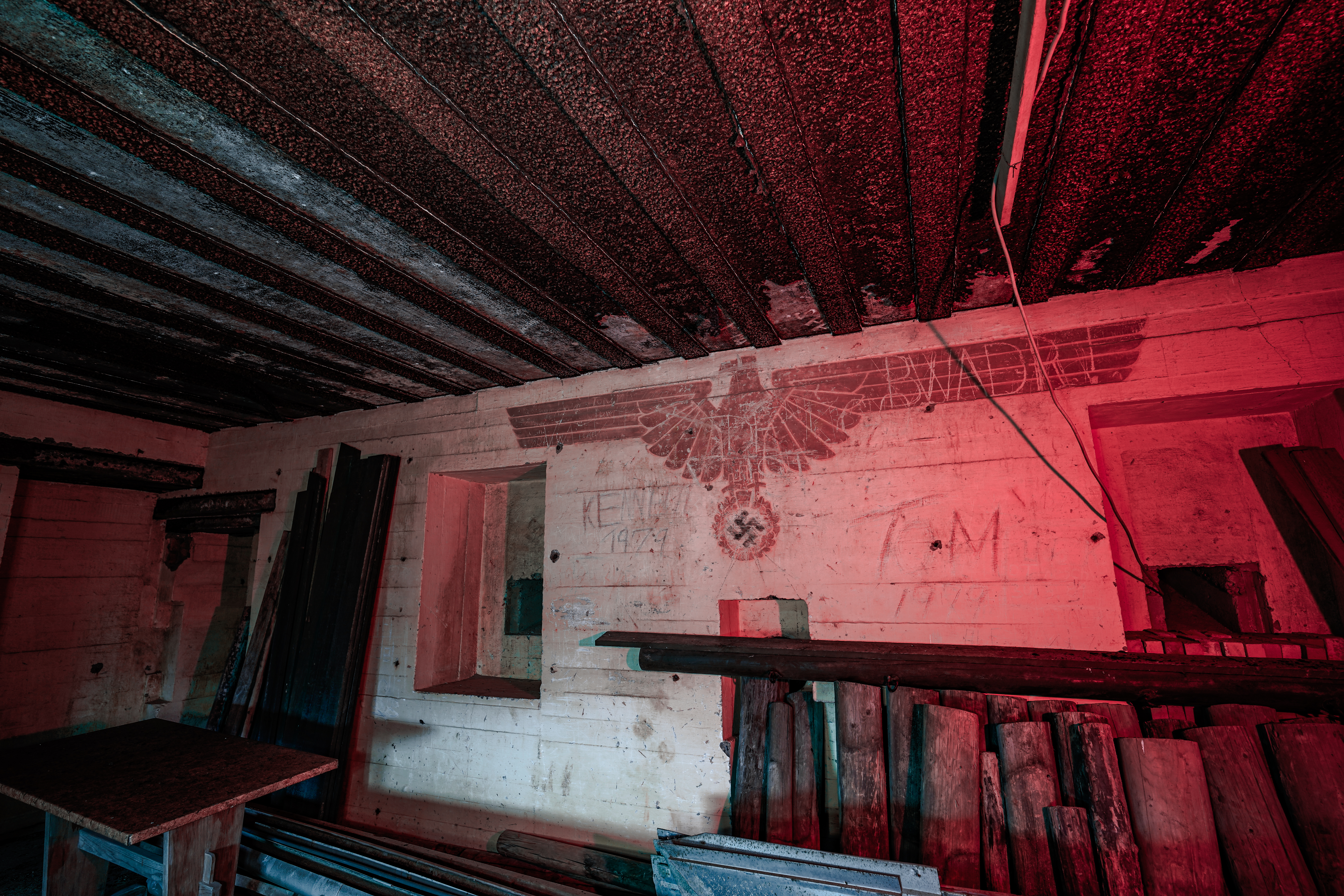 An abandoned room with a table and stacks of wooden planks, featuring a faded insignia on the wall under a red light.