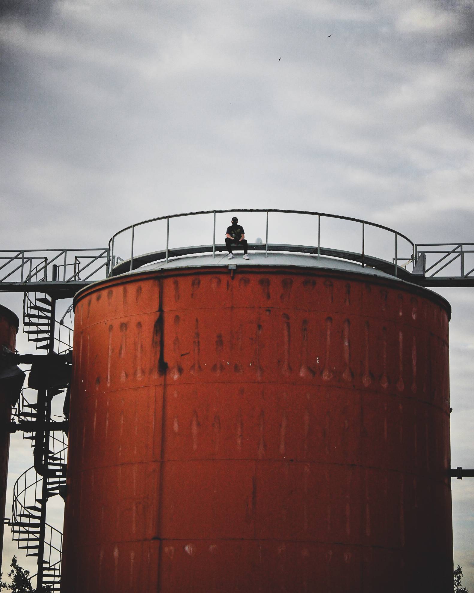 A person sitting on top of a large orange cylindrical tank, with a spiral staircase visible on the left and a cloudy sky in the background.
