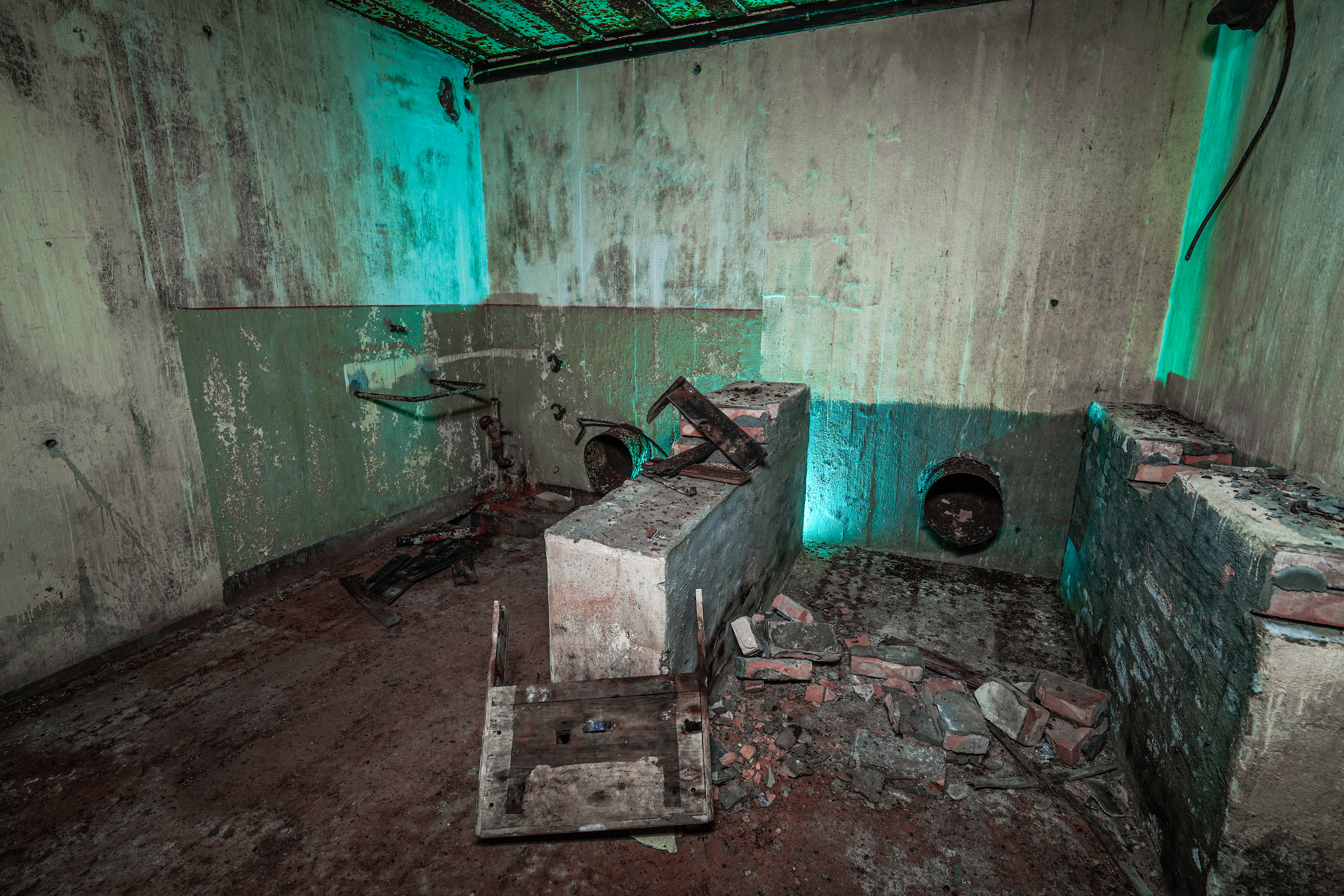 An abandoned room with concrete blocks, scattered bricks, and pipes along the walls, illuminated by turquoise light on damp surfaces.