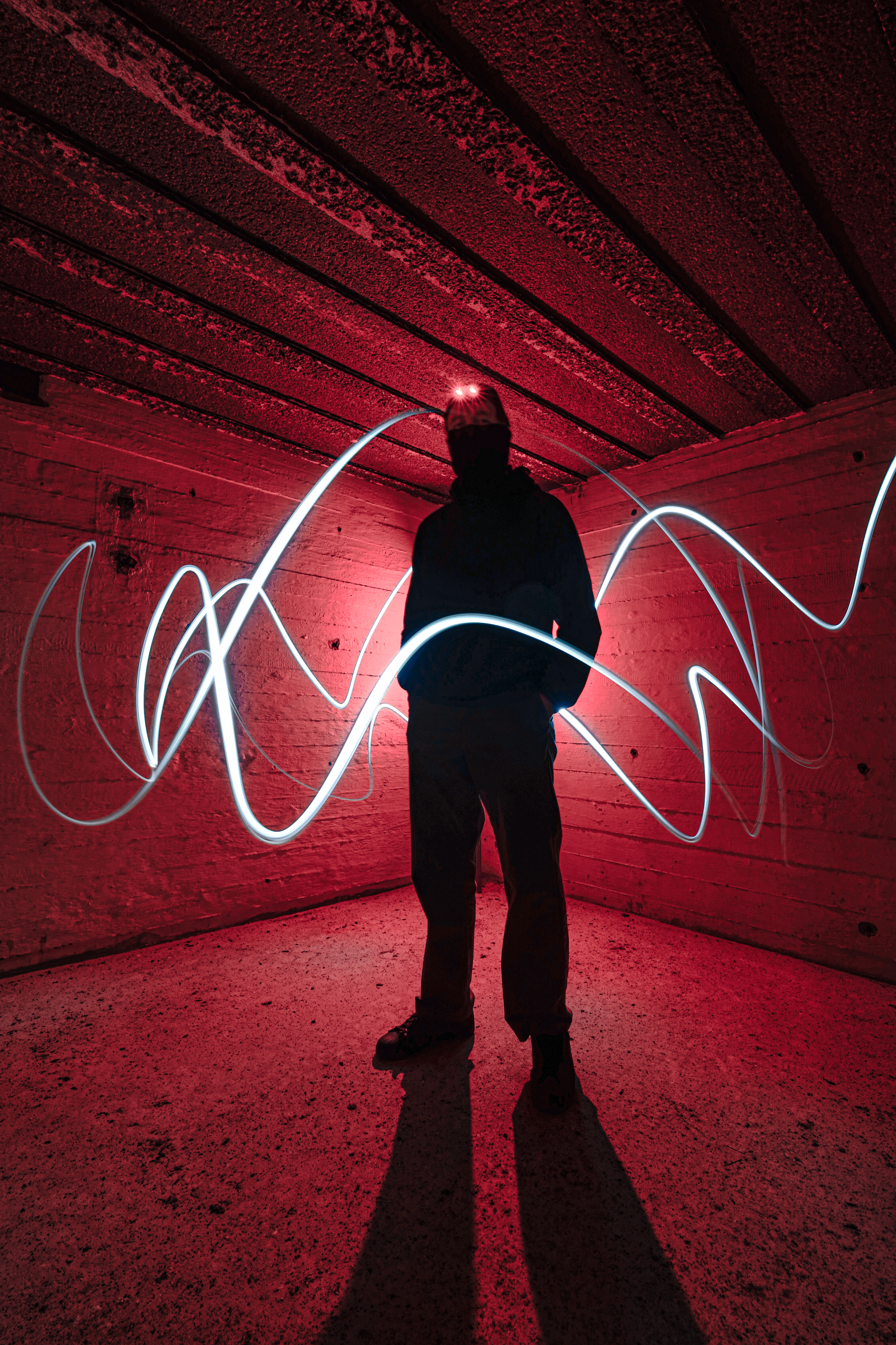 A person in dark clothing stands in an underground space illuminated by red light, with blue light trails swirling around them.