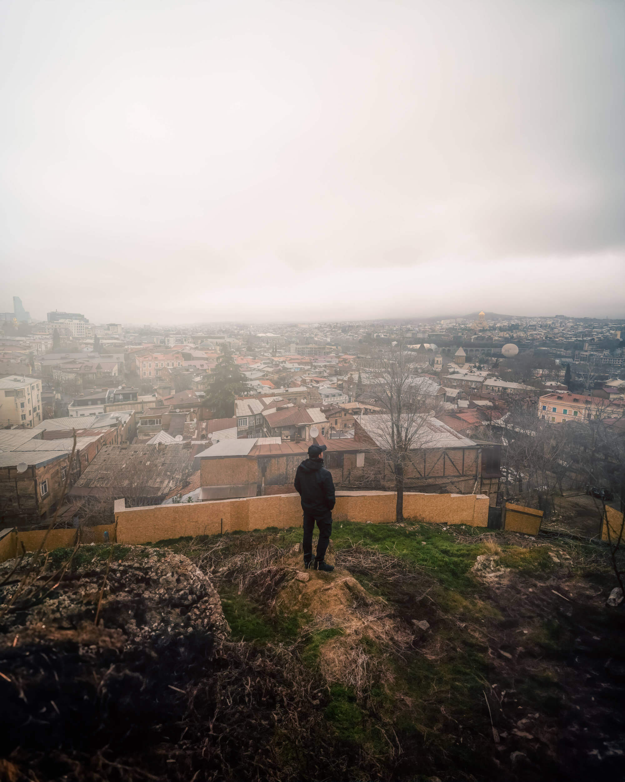 A person in dark clothing stands on a grassy hilltop, looking out over a foggy cityscape filled with rooftops and trees. The sky is overcast, adding to the subdued and contemplative mood of the scene.