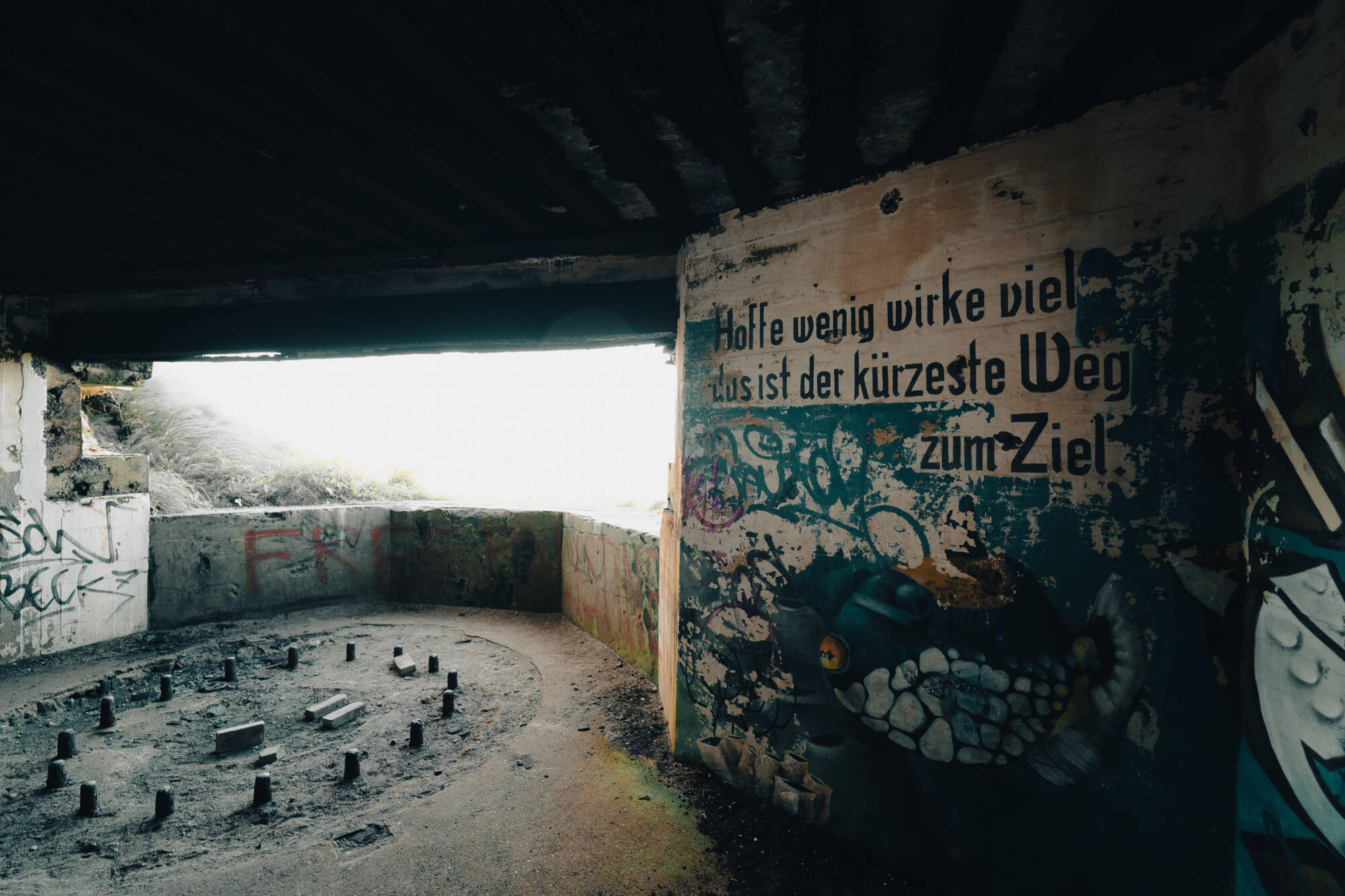 Interior of a dilapidated structure with graffiti on the walls, a circular arrangement of blocks on a dusty floor, and light streaming in from an opening.