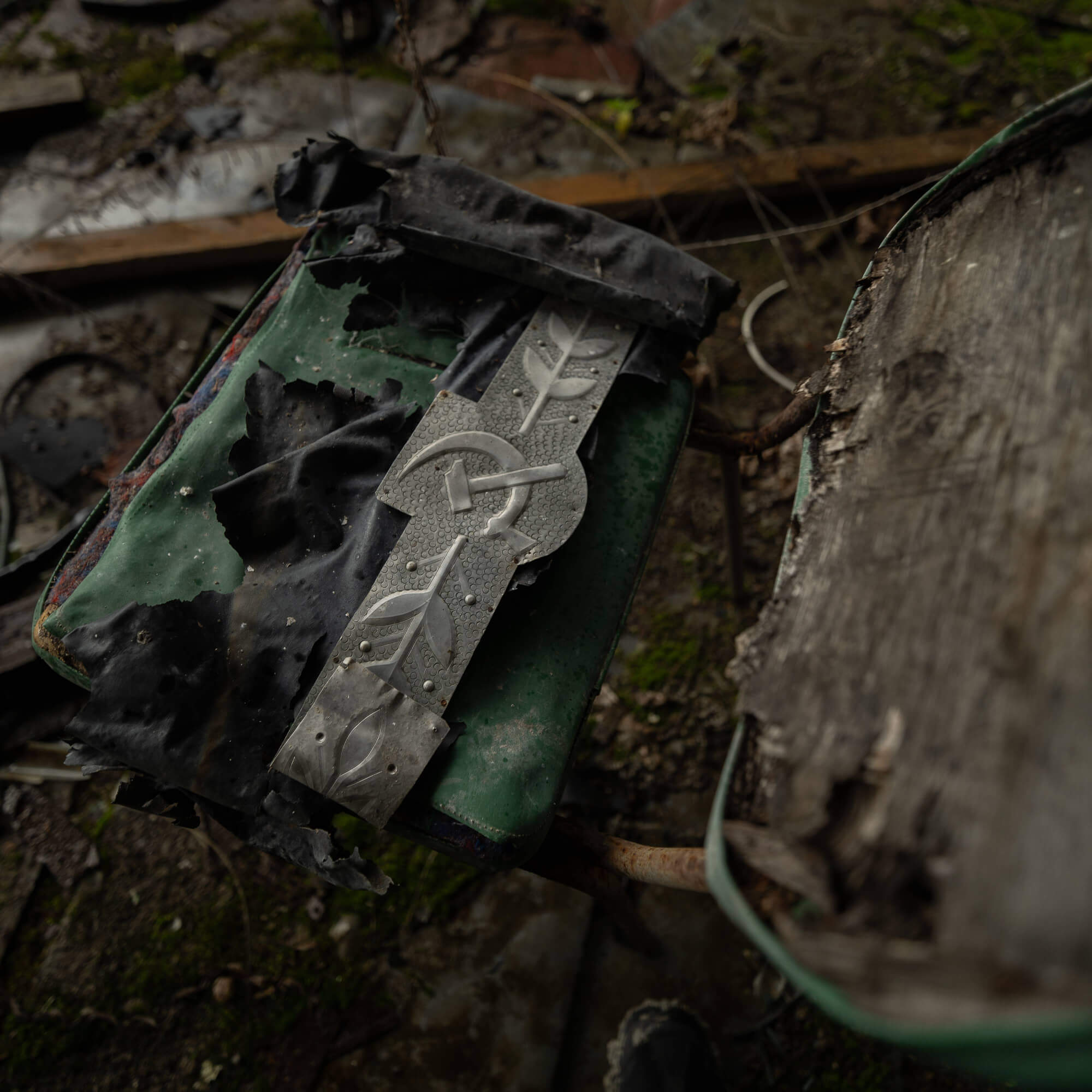 A close-up of a worn green chair with a decorative metallic belt buckle featuring leaf patterns. The chair shows signs of wear with peeling paint and crumpled black fabric nearby, set in a cluttered, decaying environment.