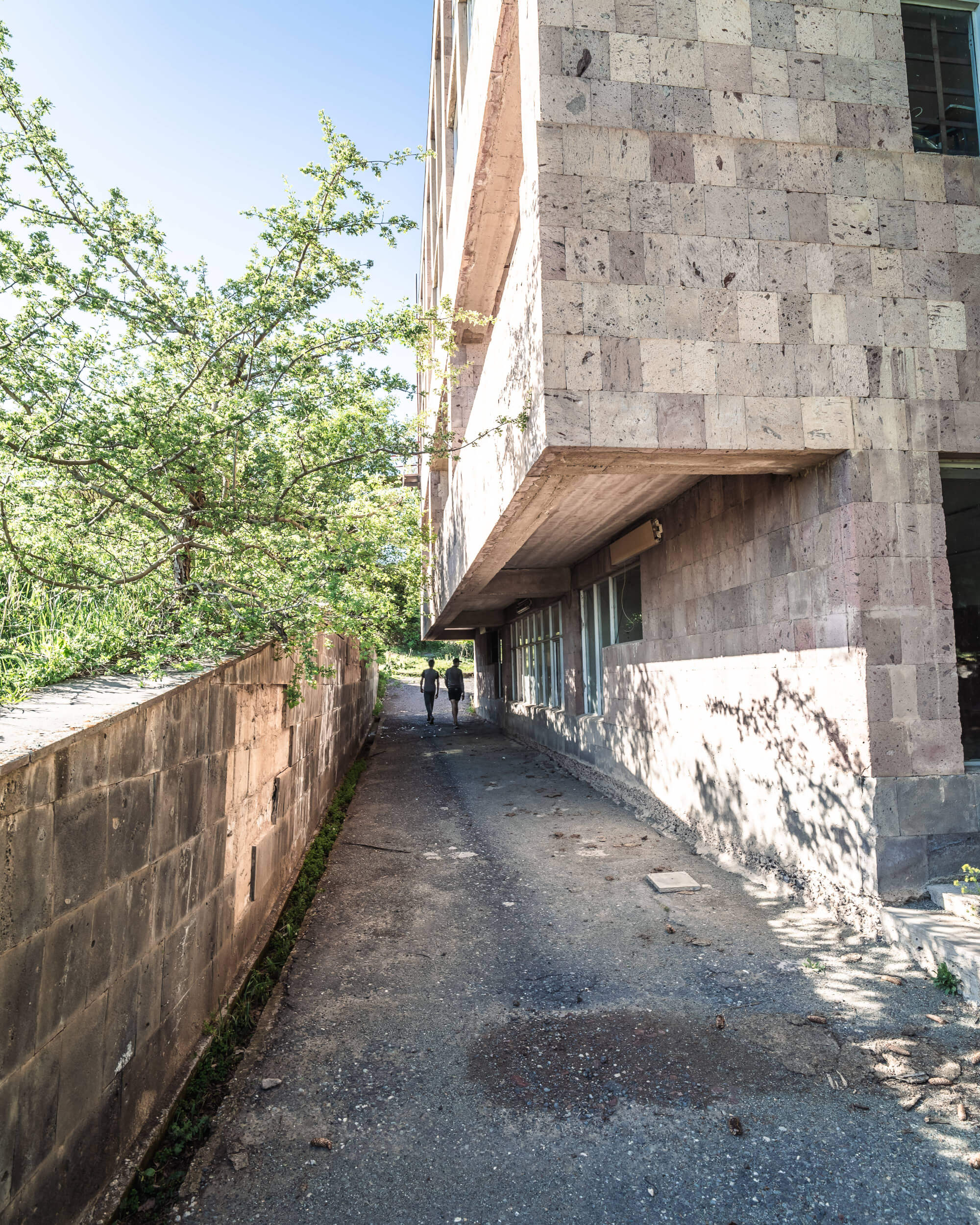 A narrow walkway alongside a textured building with two people walking. Lush greenery on one side and a stone wall on the other, under a clear blue sky.