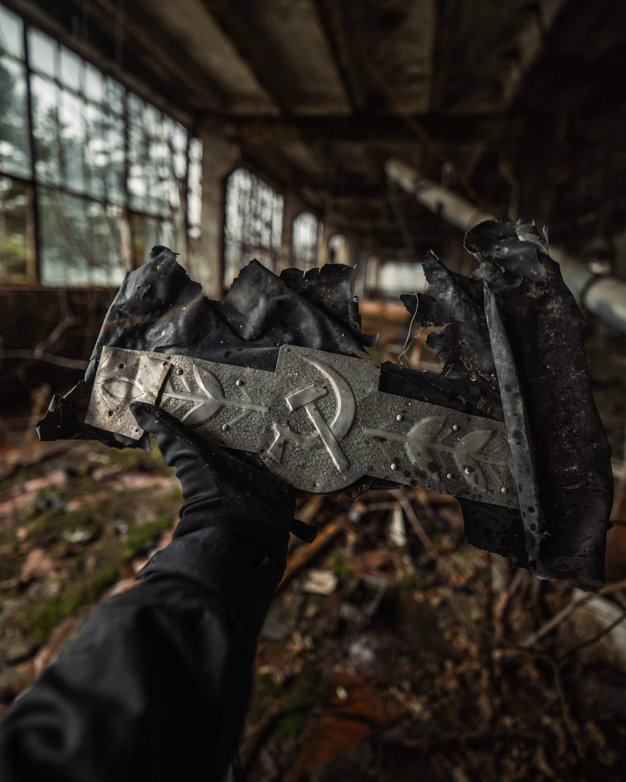 A hand in a black glove holding a worn, tattered metal object with symbols, set in a dilapidated building with broken windows and scattered debris.