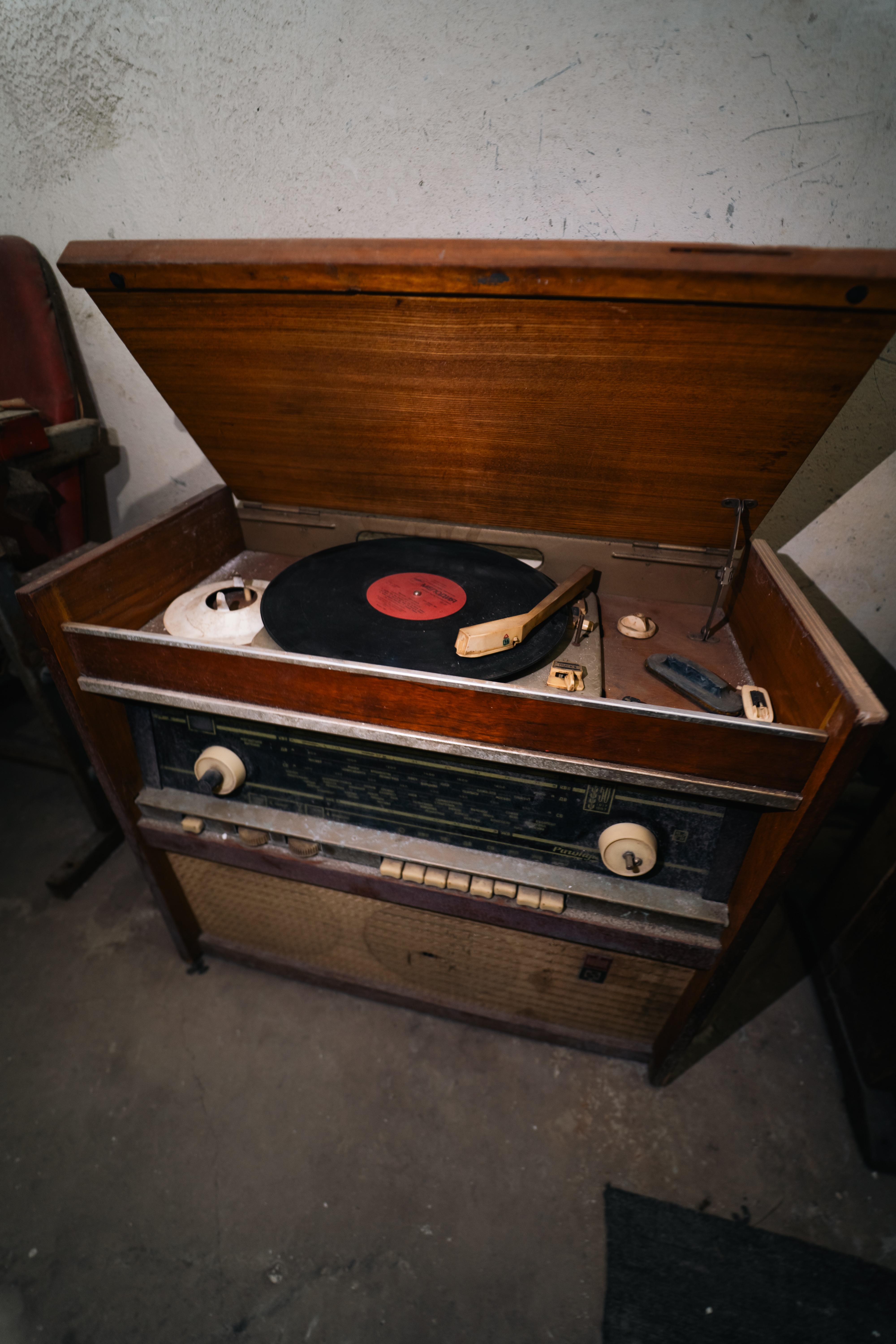 A vintage wooden record player with a black vinyl record on it, situated in a dusty room. The lid is open, revealing the record and a tonearm, with buttons and knobs visible on the front panel.