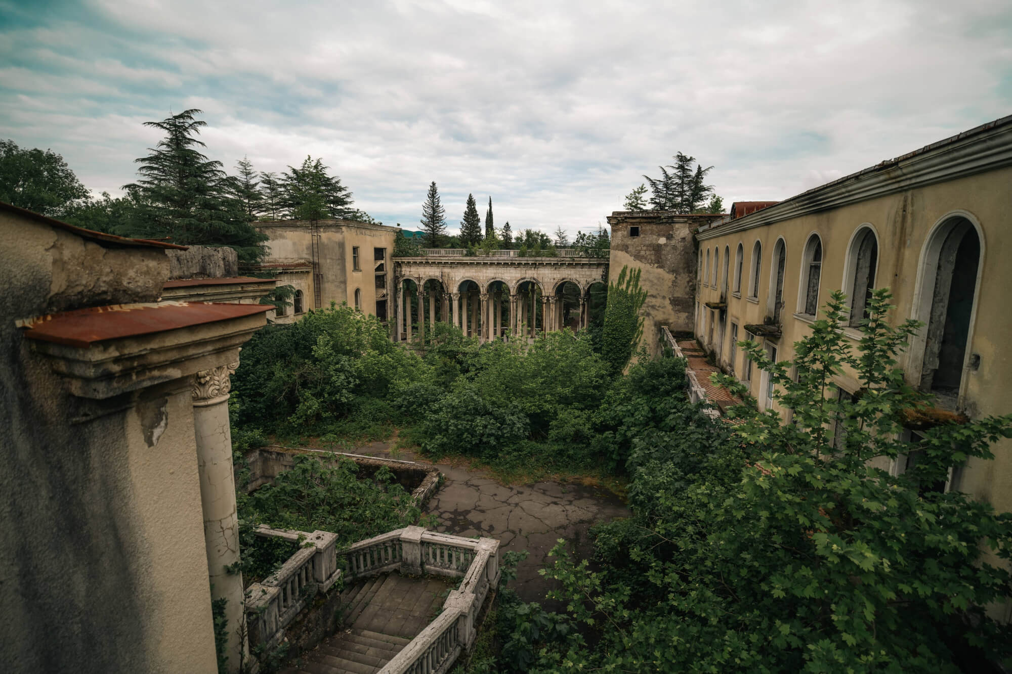 A view of an abandoned building surrounded by lush greenery, featuring crumbling arches and a staircase partially hidden by plants, under an overcast sky.
