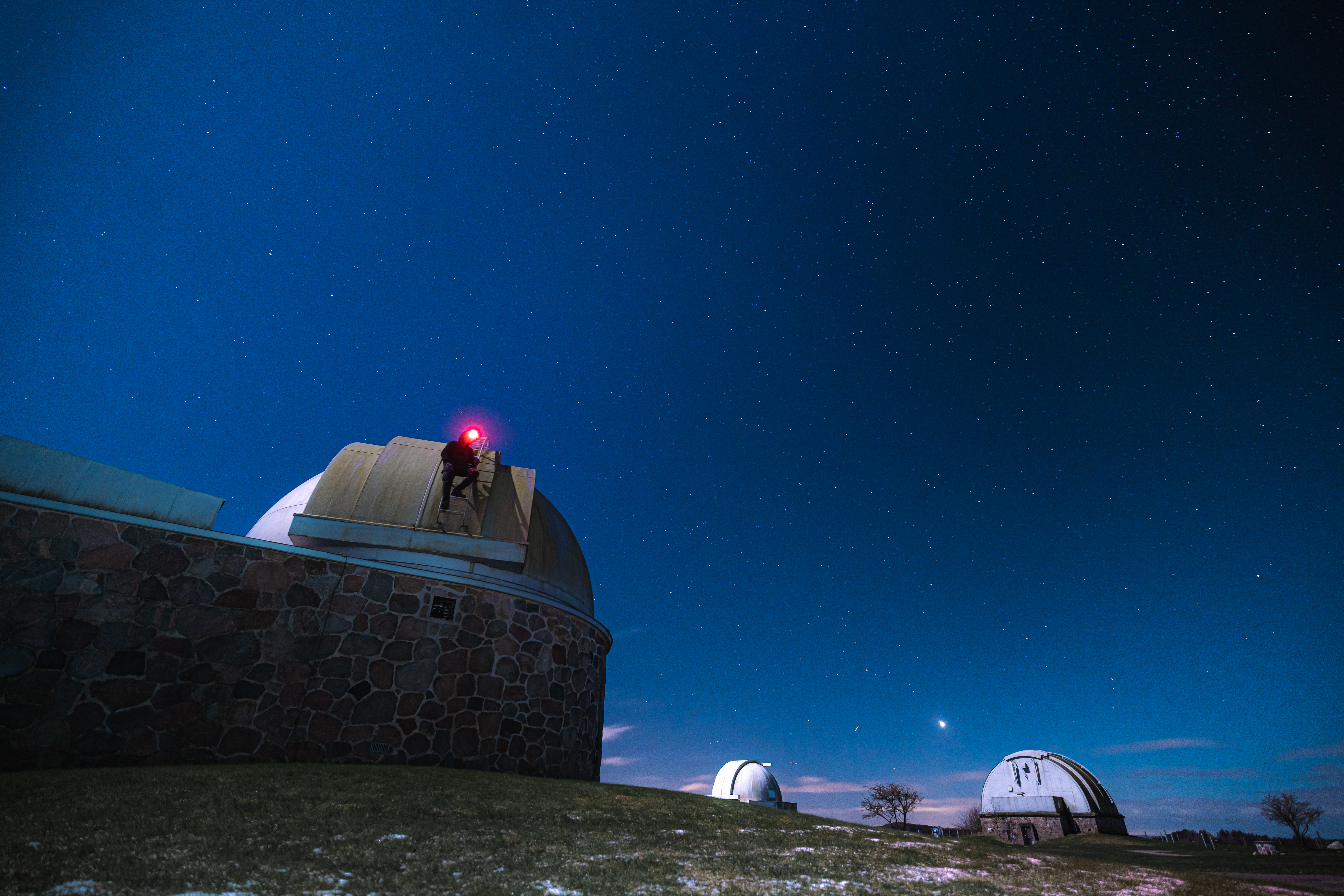 A person climbing a ladder on an observatory with a red light on top, under a star-filled blue sky. Two observatory domes are visible in the background, with a grassy foreground.