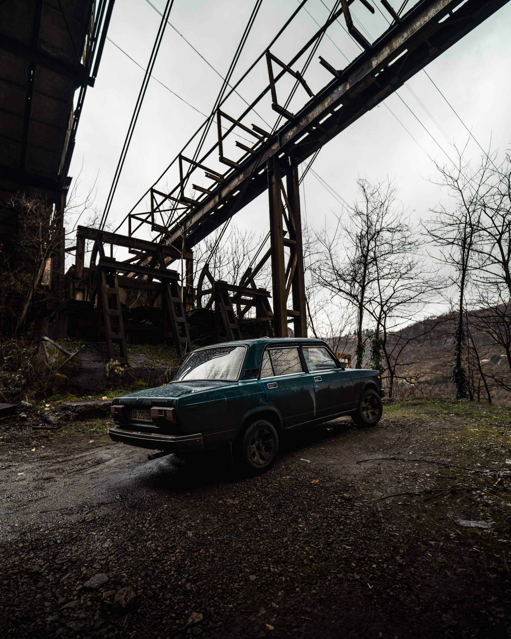 A blue car parked on a gravel path near a rusted industrial structure, surrounded by bare trees and distant hills under an overcast sky.