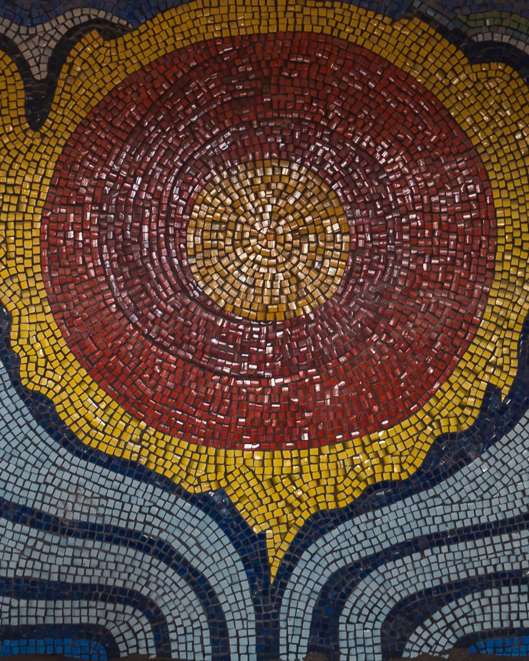 Close-up of a colorful mosaic featuring a circular sun design in reds and yellows, surrounded by curved patterns in various shades of blue.