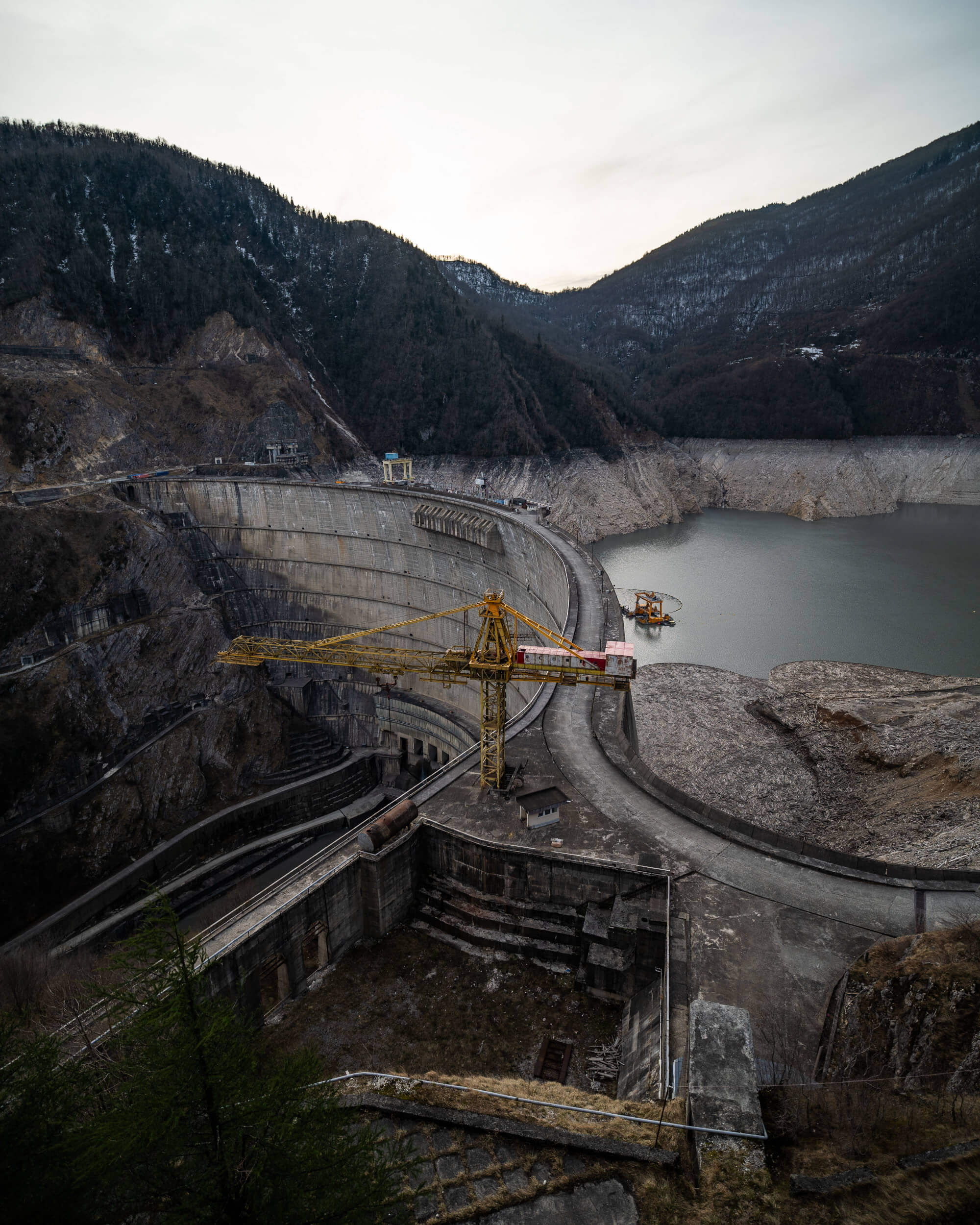 A large concrete dam with a yellow crane on top, surrounded by mountains and a calm reservoir. The sky is overcast, and the landscape features rocky terrain and sparse vegetation.