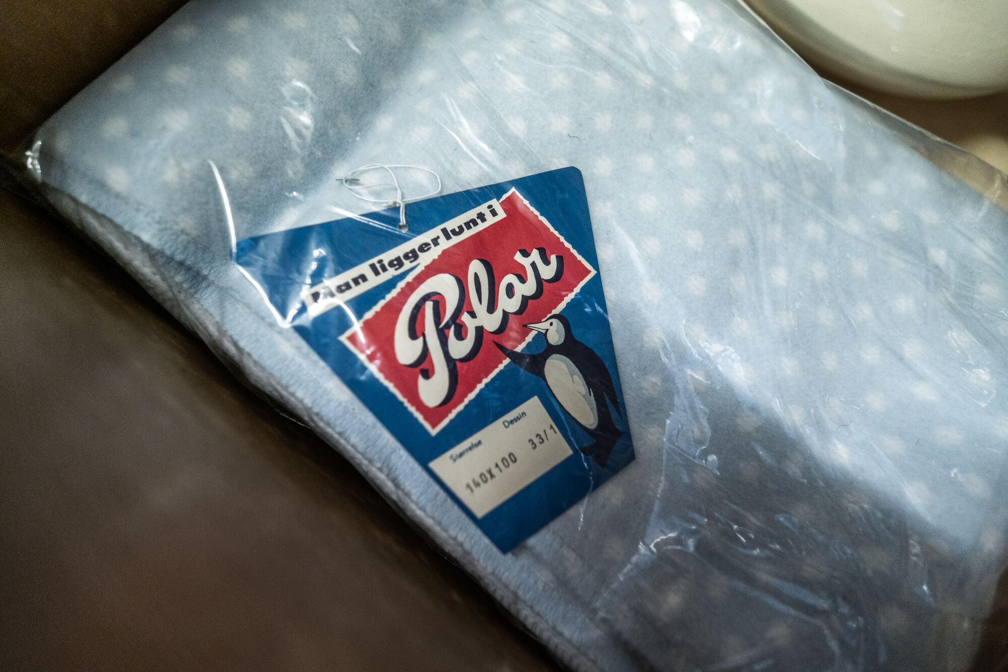 A close-up of a blue polka dot blanket wrapped in plastic with a colorful label featuring a cartoon penguin and text, placed in a cardboard box.