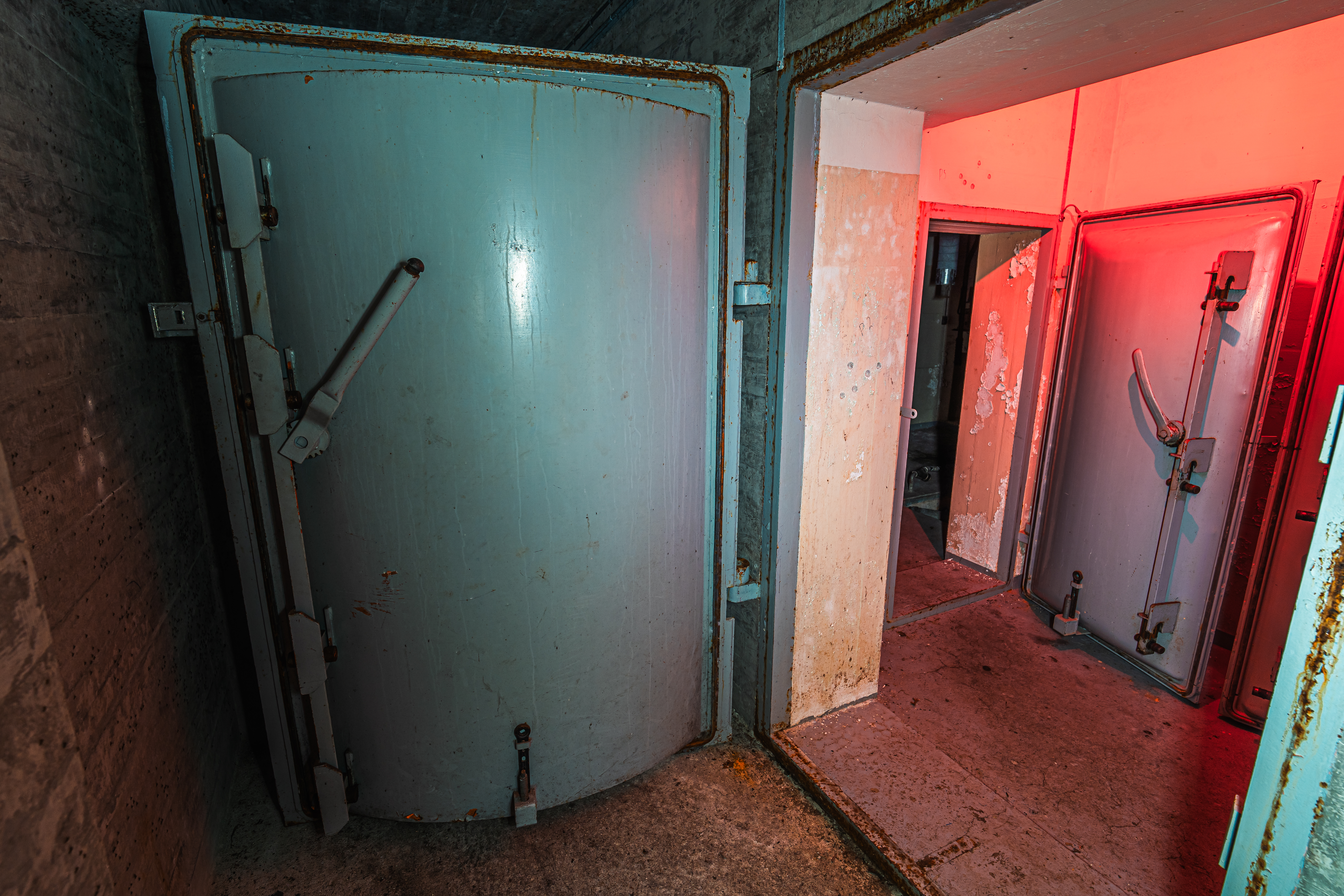 An abandoned room with two large, rusted metal doors; one blue and one gray, with peeling walls and a red light illuminating the space.