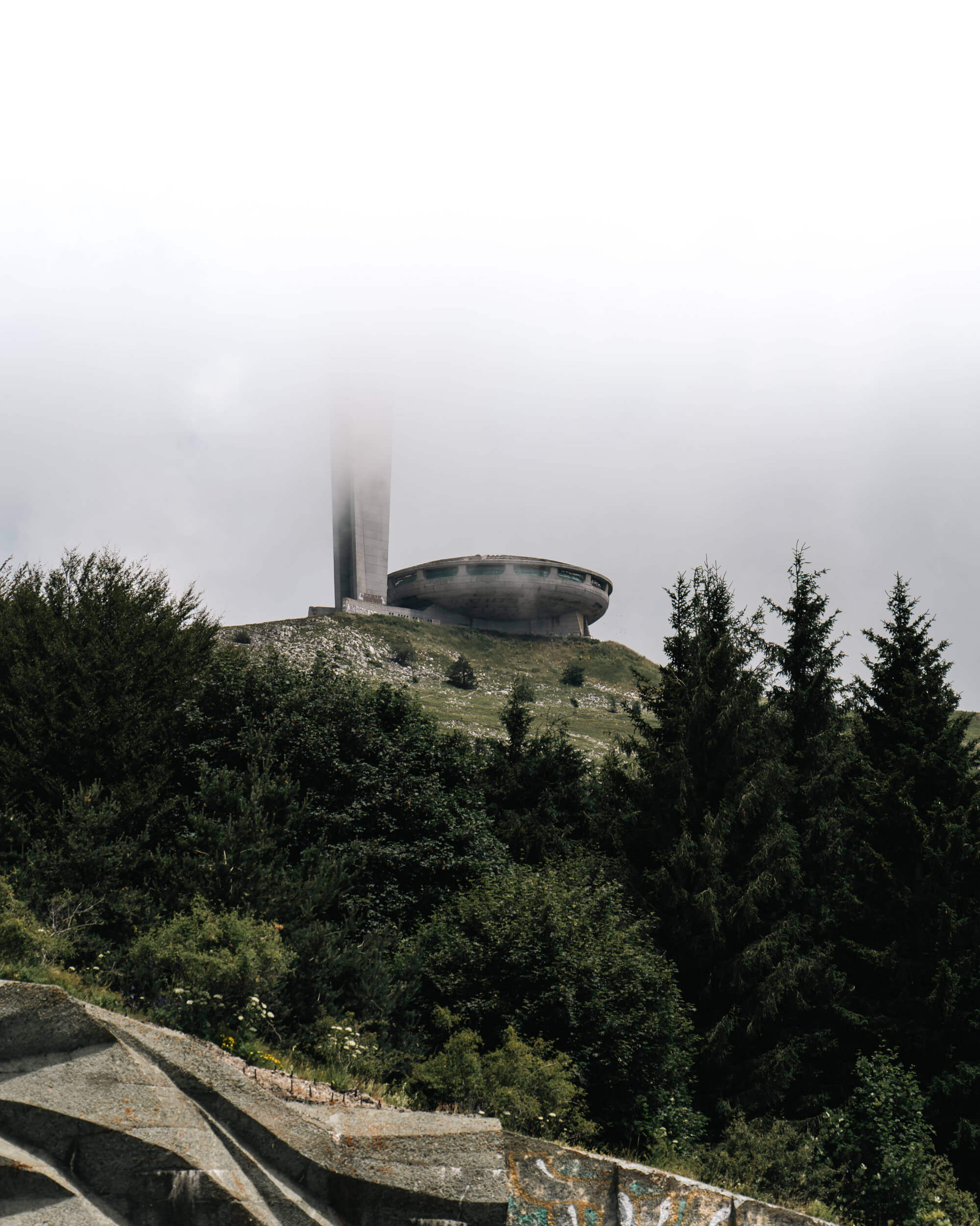 A fog-covered hill with a large, circular, abandoned structure at the top, surrounded by dense trees and greenery. The foreground features rugged stone textures.