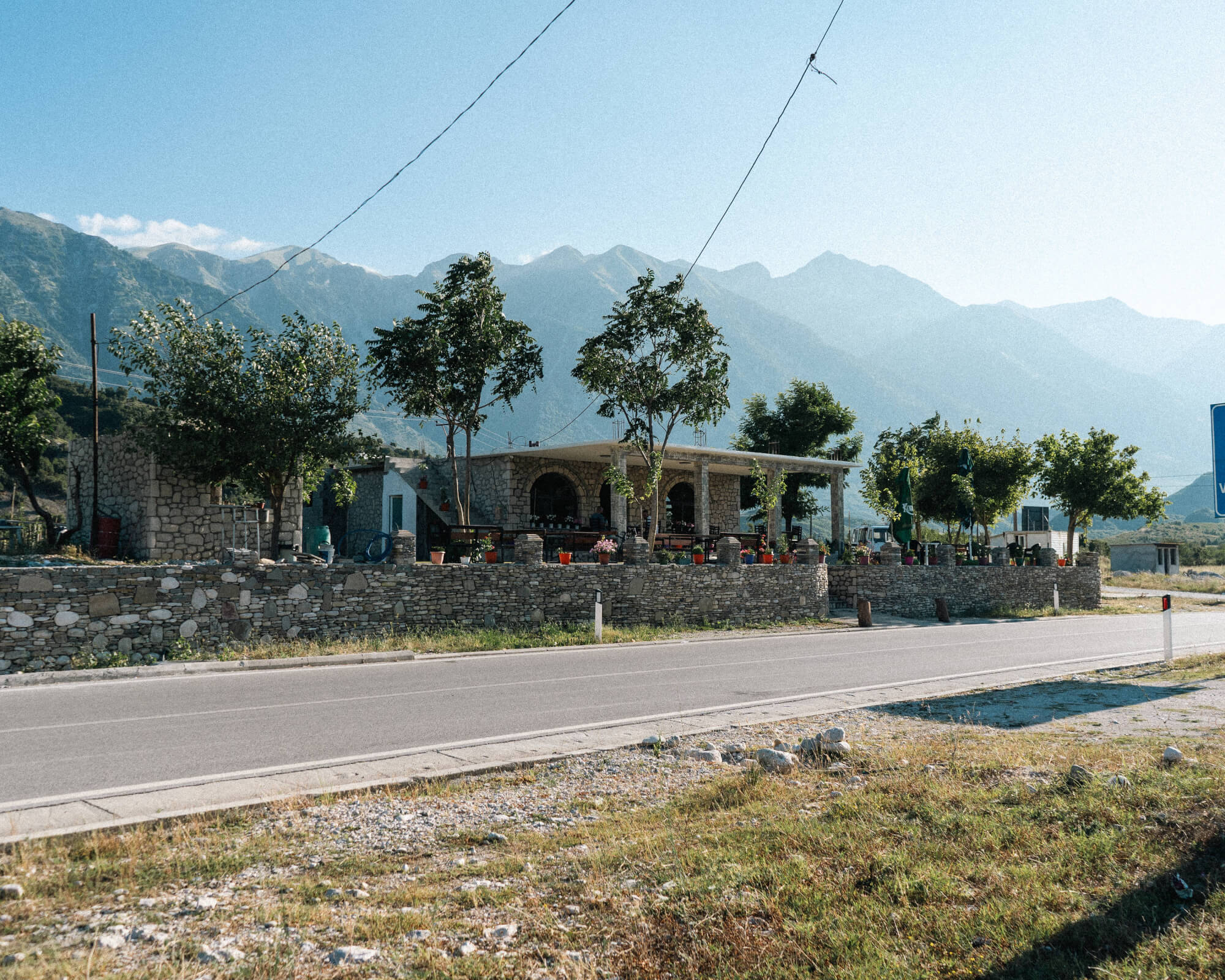 A stone house with a porch surrounded by trees, located near a road, with mountains in the background and a clear blue sky.