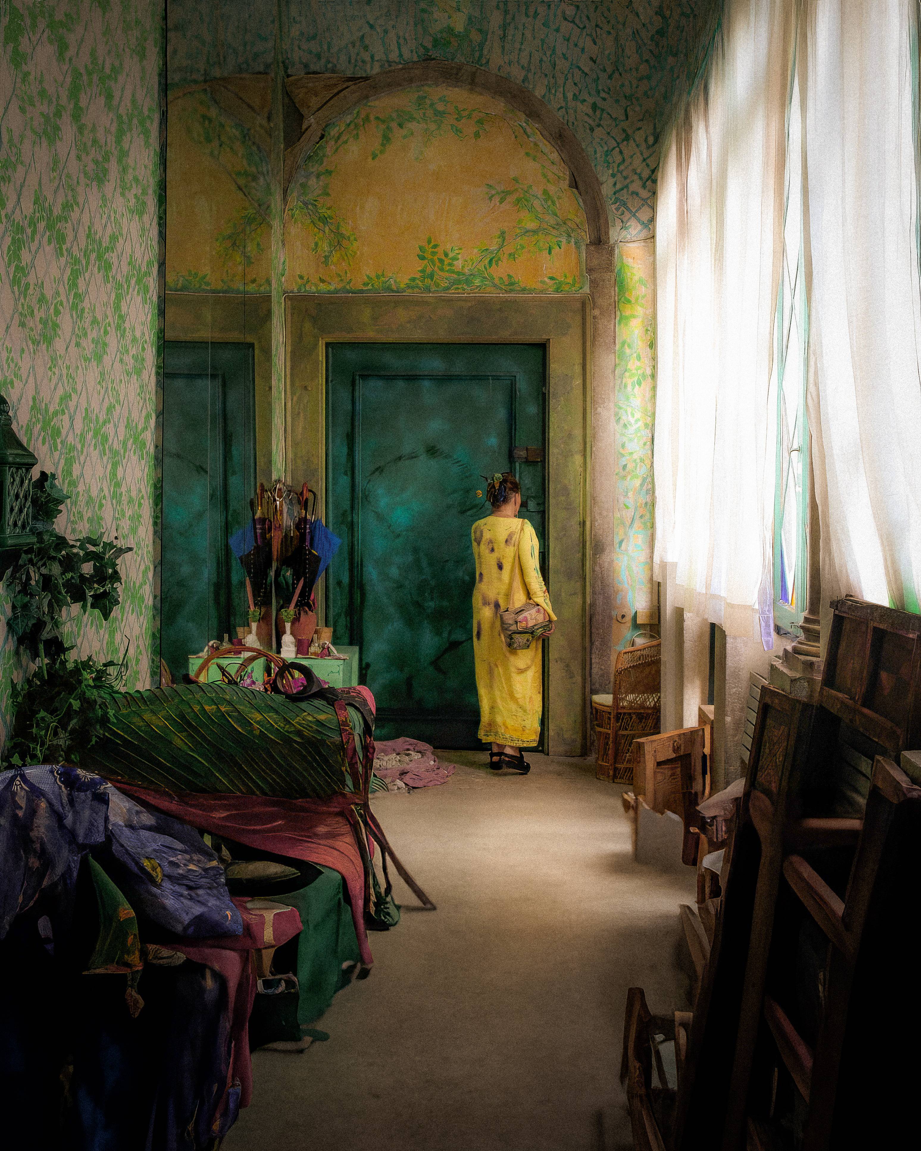A person in a yellow dress standing at a doorway in a colorful room with floral wallpaper. Soft light comes through white curtains, illuminating various textiles and umbrellas in the space.