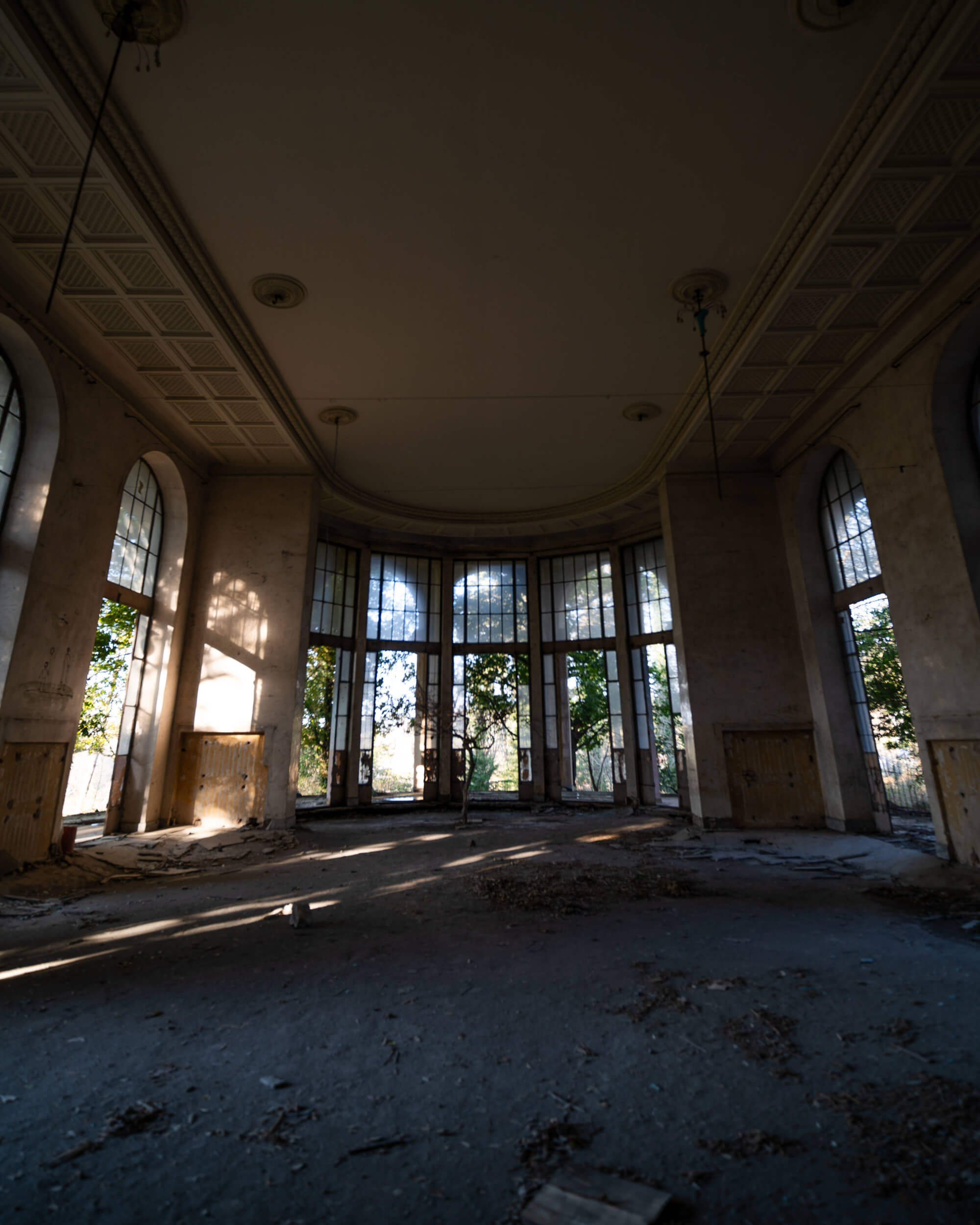 An abandoned room with large arched windows allowing sunlight to stream in, highlighting dust and debris on the floor. There are hints of greenery visible outside, indicating nature's encroachment into the space.