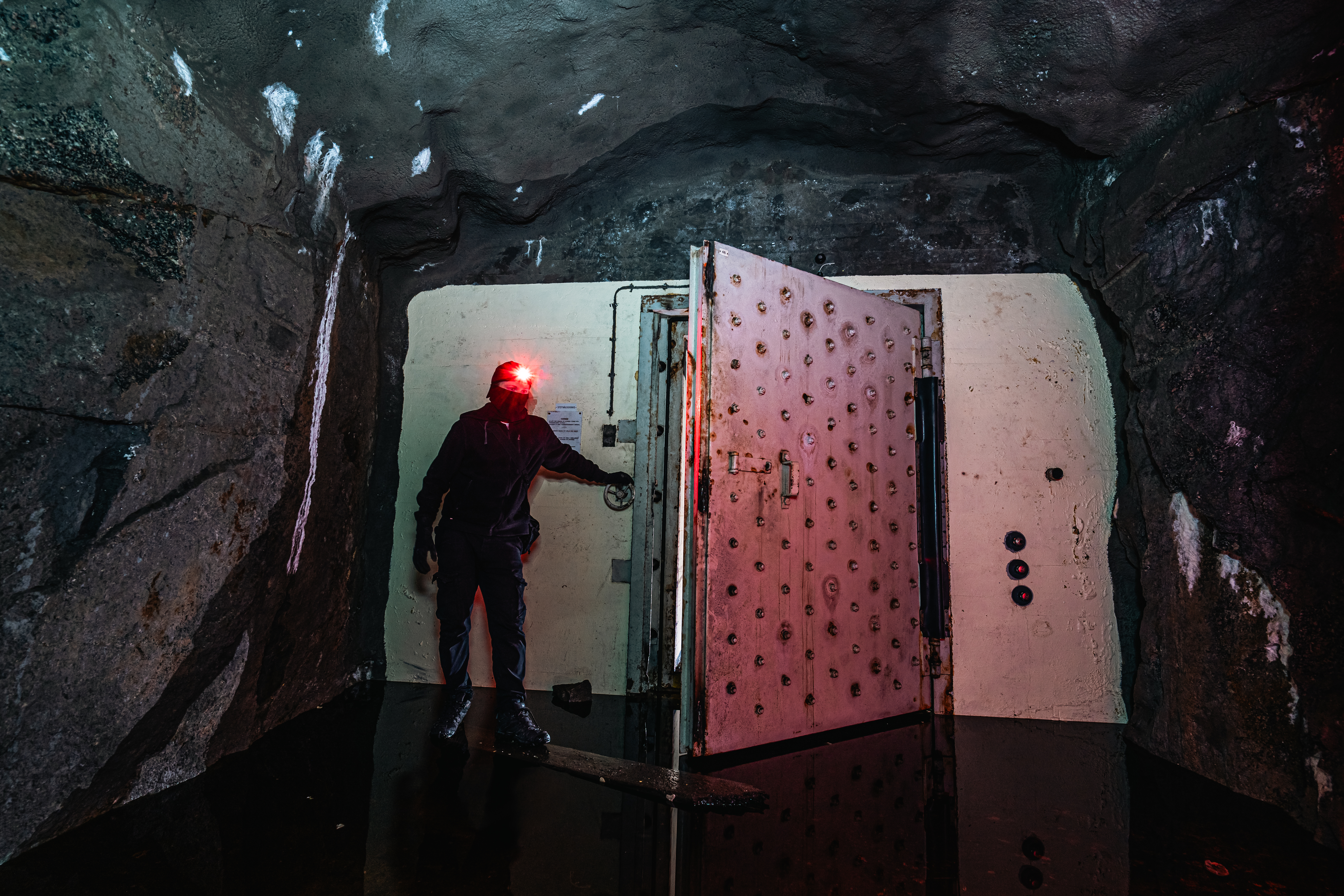 A dark cavern with a person wearing a black outfit and a red headlamp standing in front of a large, rusty door. The ground is covered in water, reflecting light. The walls are rocky and rugged.