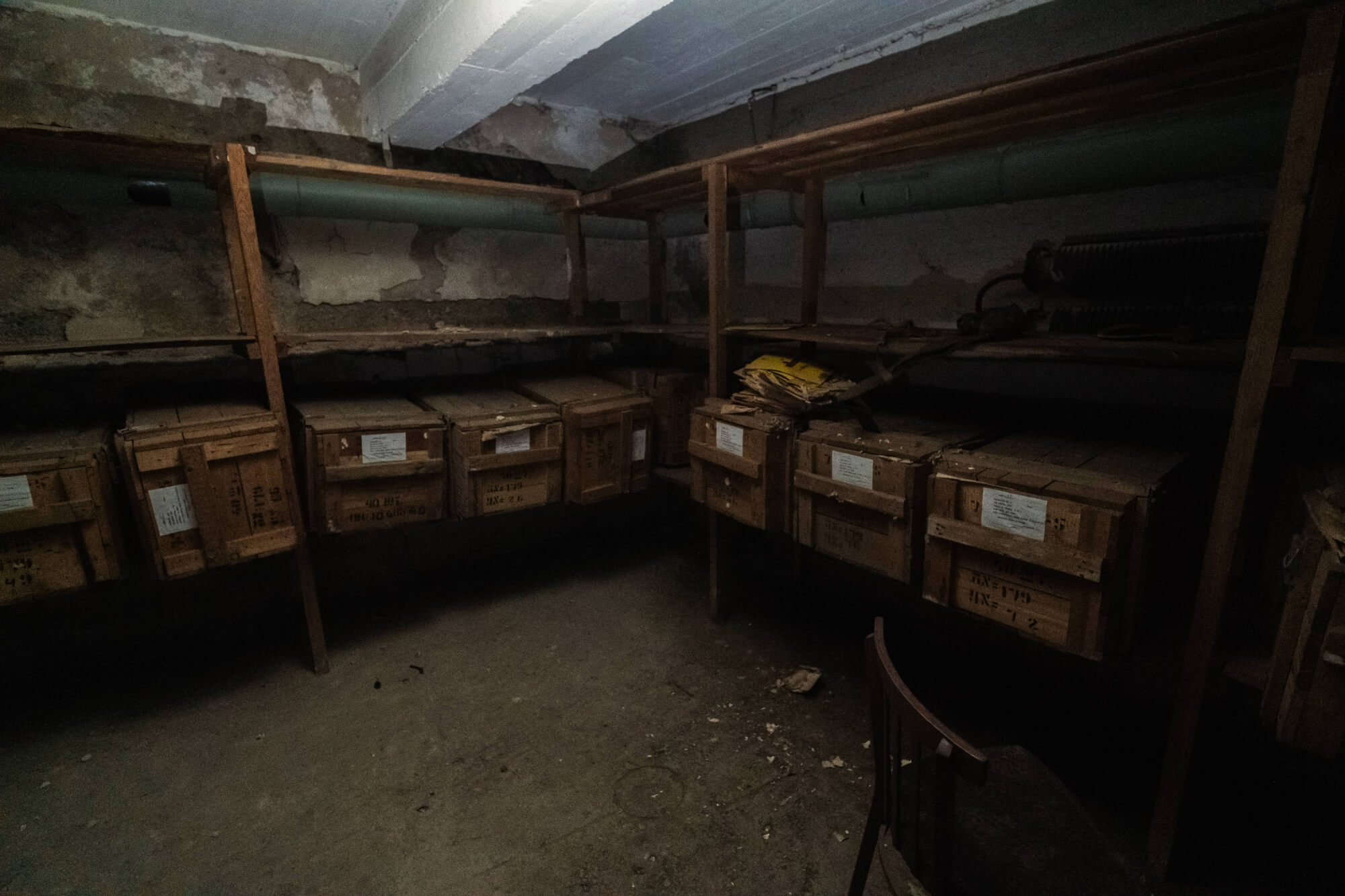 A dimly lit storage room with wooden crates on shelves, dust particles in the air, and a chair in the corner.
