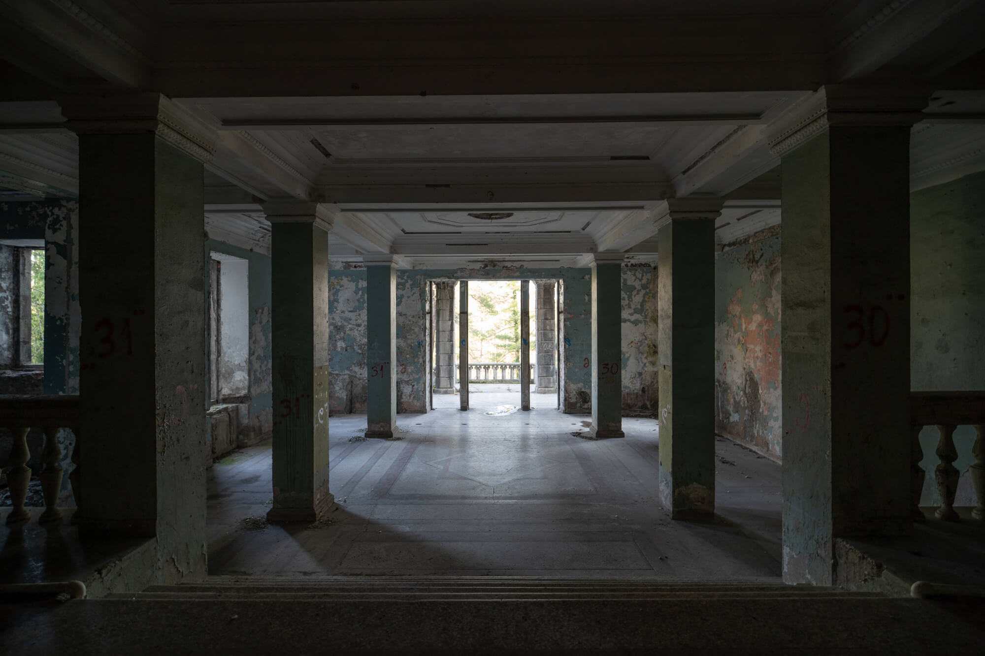An empty, dilapidated room with peeling paint, columns, and two open doors in the distance leading to natural light.