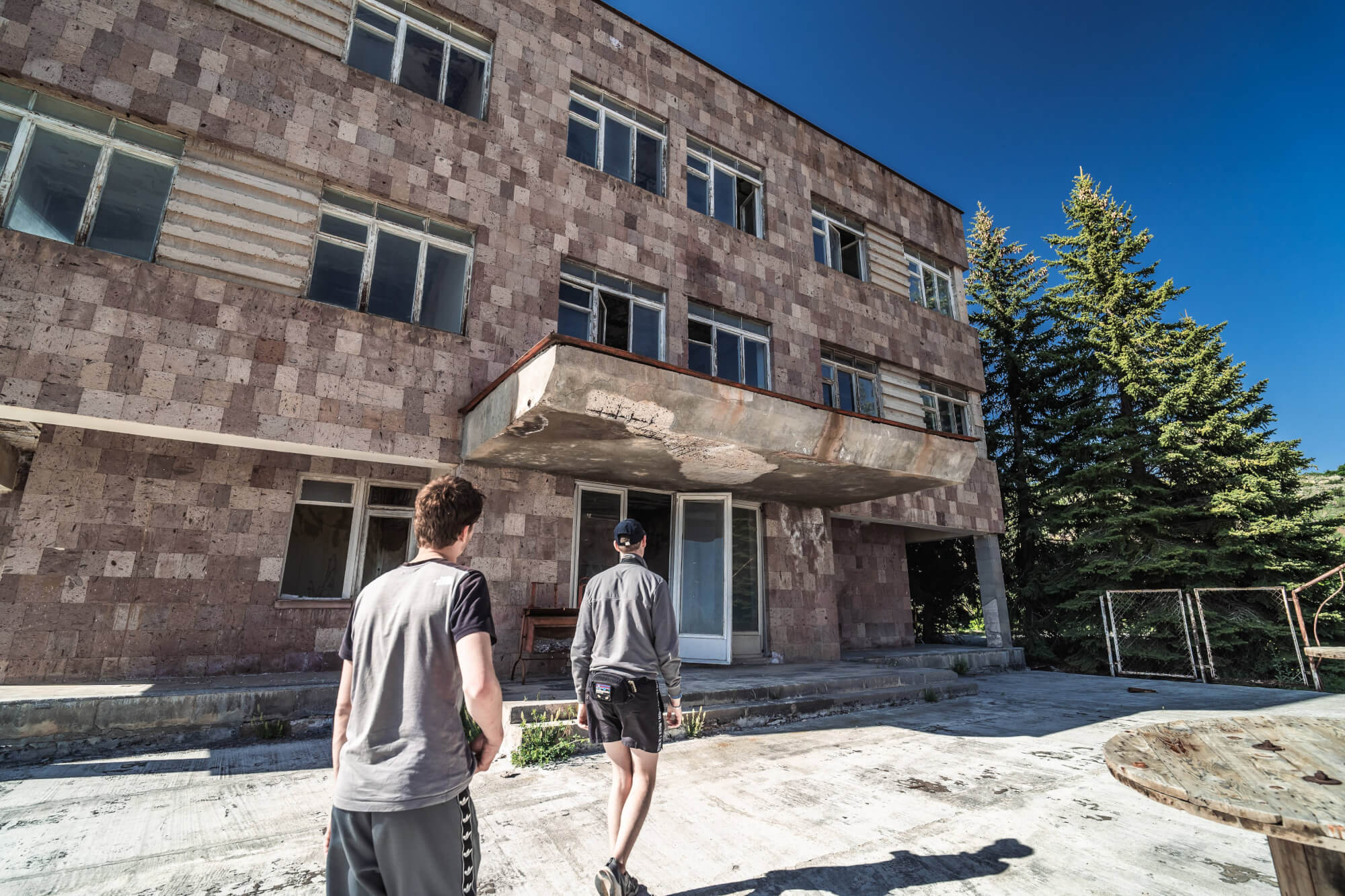 Two people walking towards an old, abandoned building with a textured, brown facade and overhanging balcony, surrounded by greenery and under a clear blue sky.