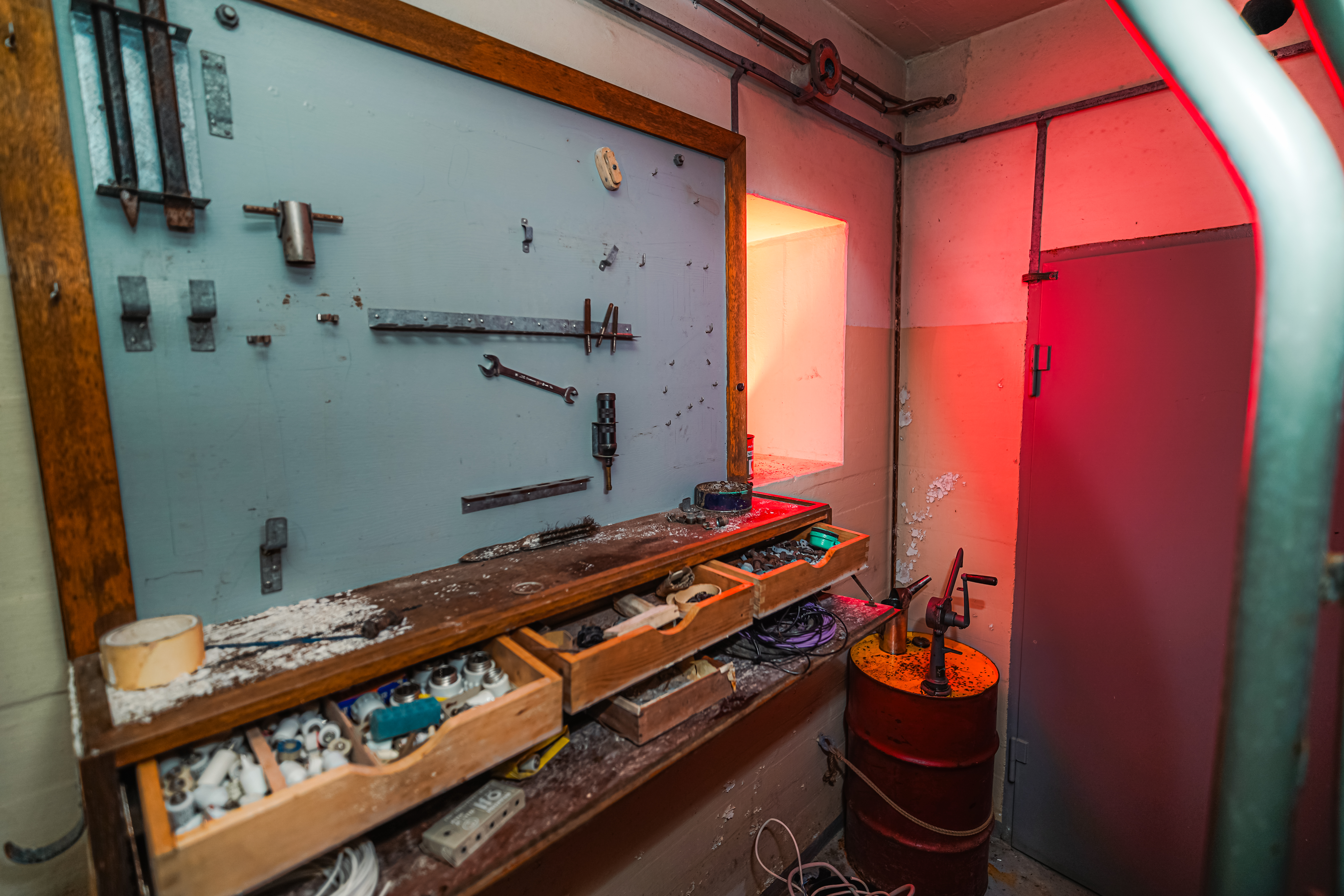 An industrial workshop with wooden shelves filled with tools, a dimly lit workspace, and a red-glowing window. The scene includes various utensils and a rusted barrel, projecting an atmosphere of nostalgia and creativity.