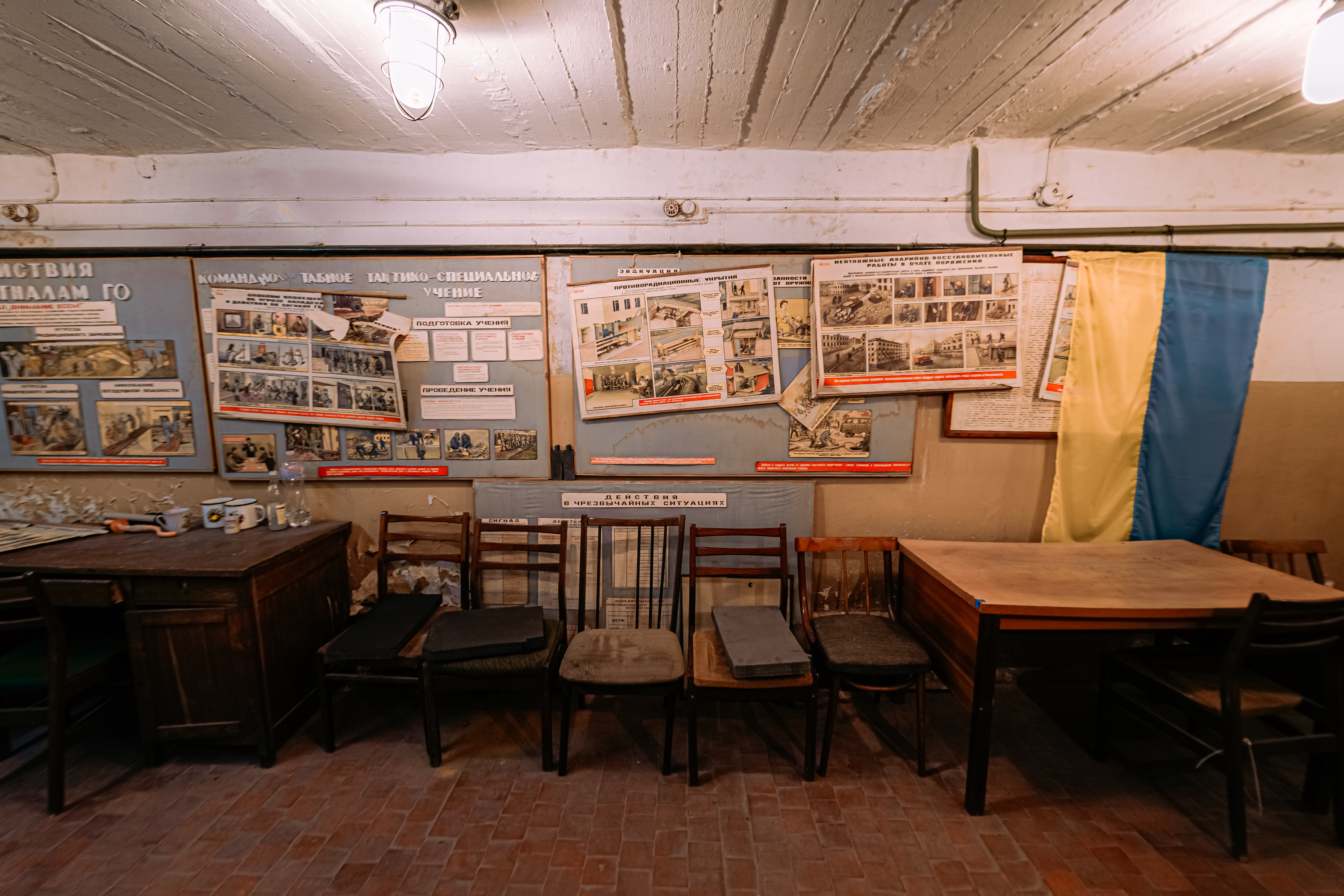 An interior space with wooden chairs and tables, adorned with educational posters on the walls. A Ukrainian flag hangs in the corner, and the room is illuminated by a single light bulb.